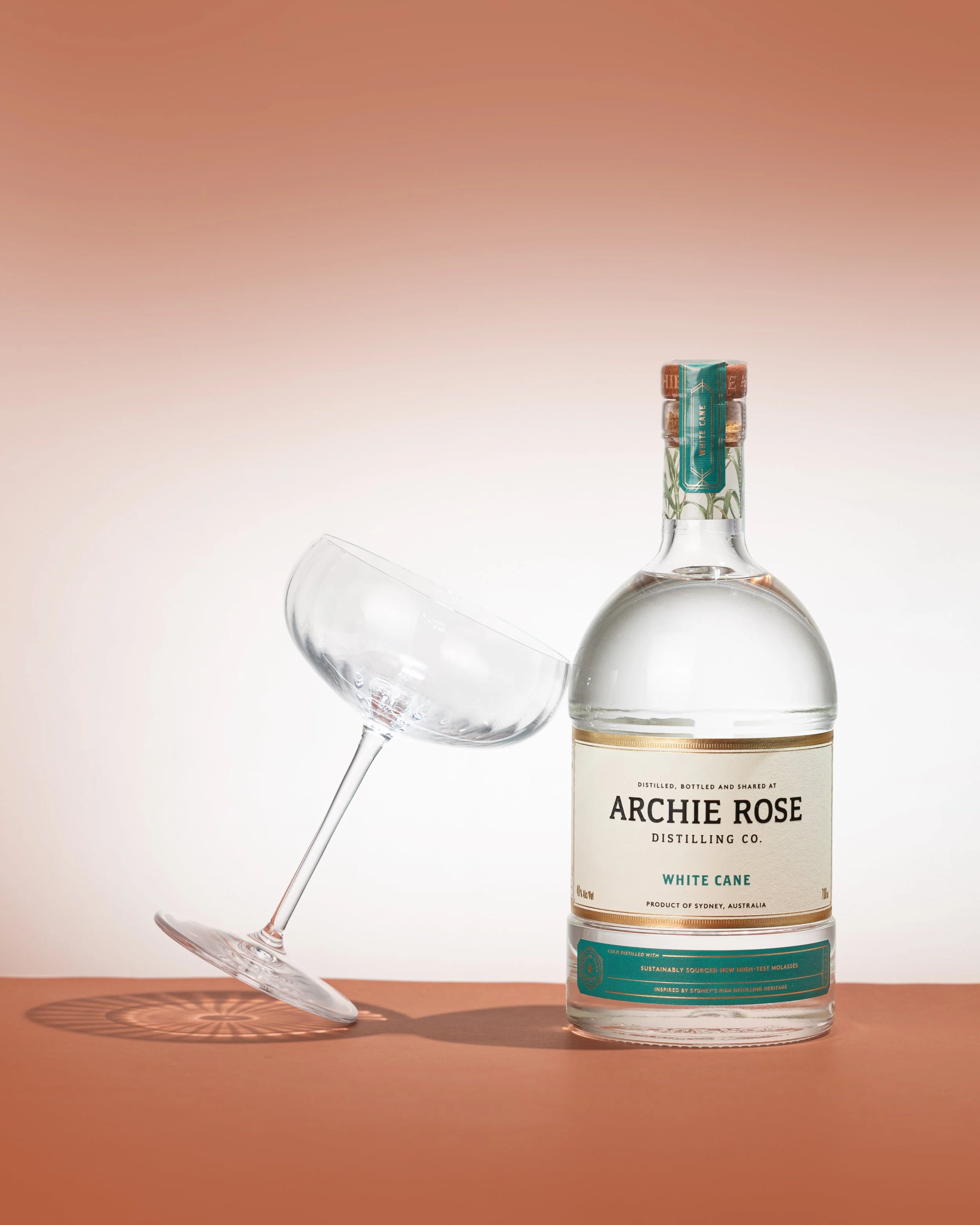 A clear glass wine bottle labeled 'Archie Rose,' 'White Cane,' and 'Distilling Co.,' with a blue and orange label, and a tilted empty wine glass leaning against it, on an orange surface with a gradient background. Photography by www.lilamarvell.com.a