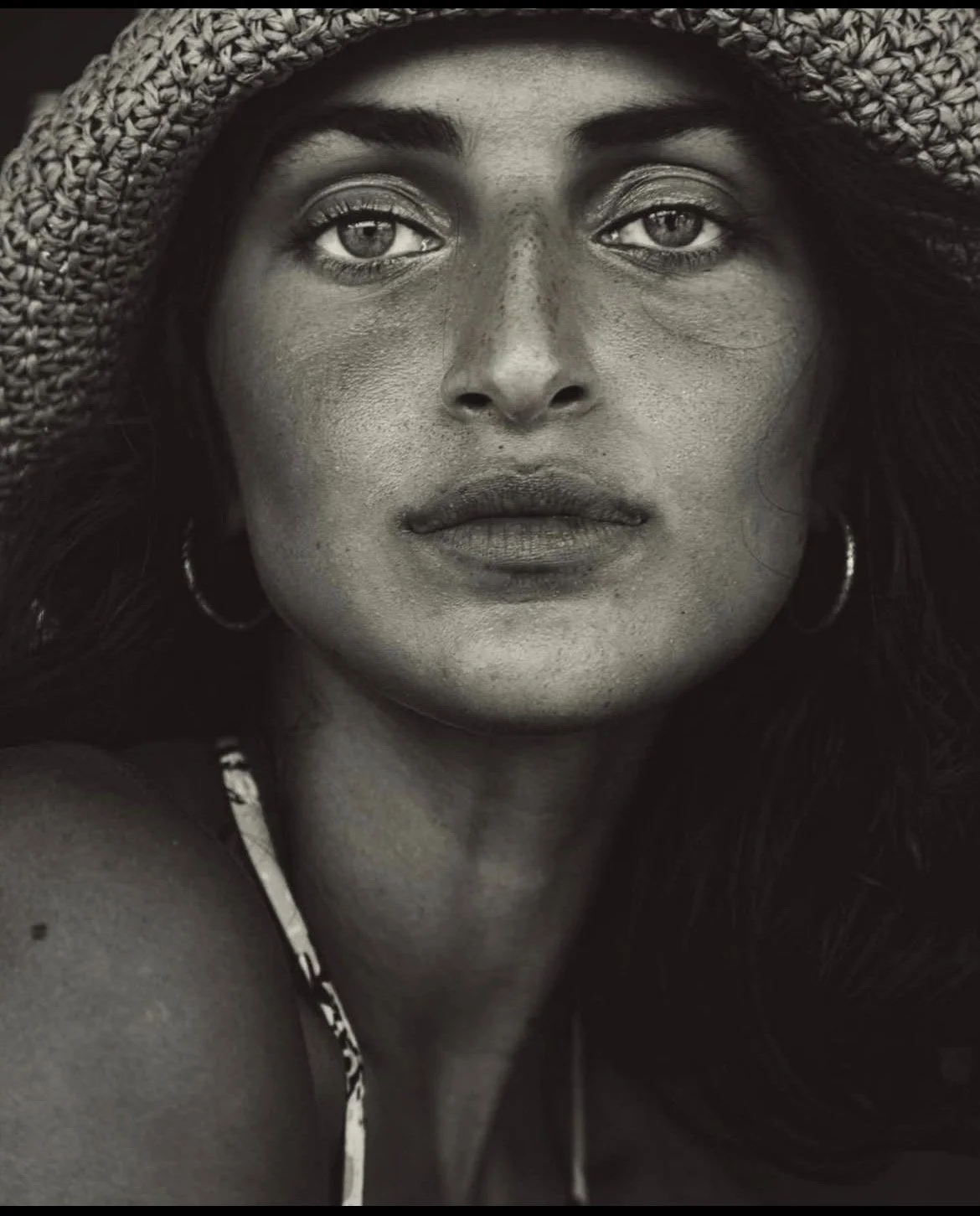 A black and white close-up portrait of a woman wearing a wide-brimmed straw hat, hoop earrings, and a patterned top or swimsuit strap, looking directly at the camera.
