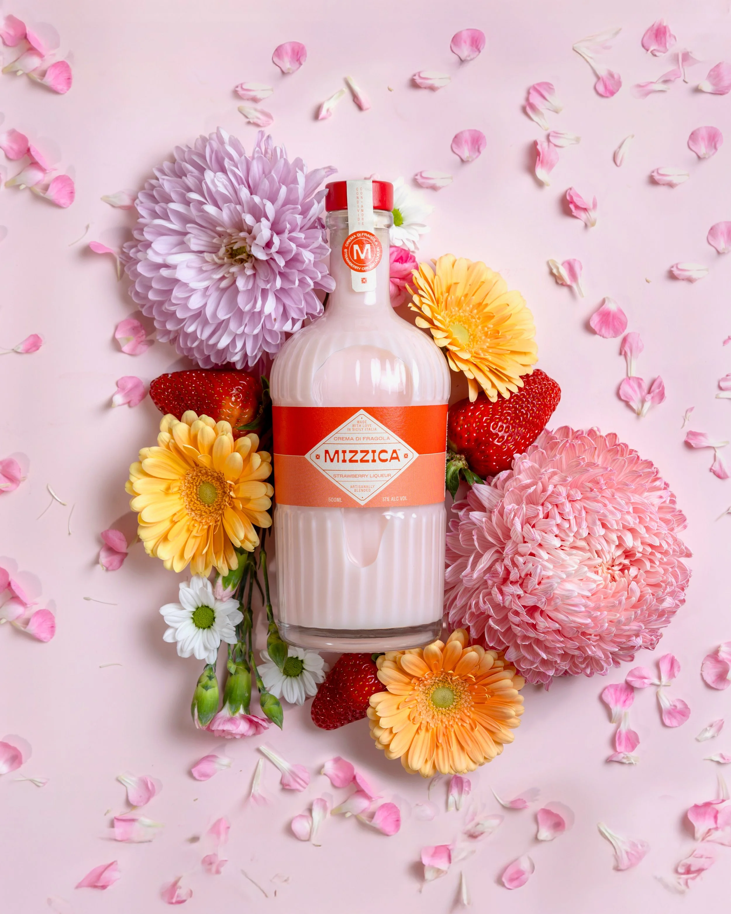 A bottle of Mizzica Strawberry Liqueur surrounded by pink and yellow flowers and scattered pink flower petals on a pink background. Photography by Lila Marvell www.lilamarvell.com.au