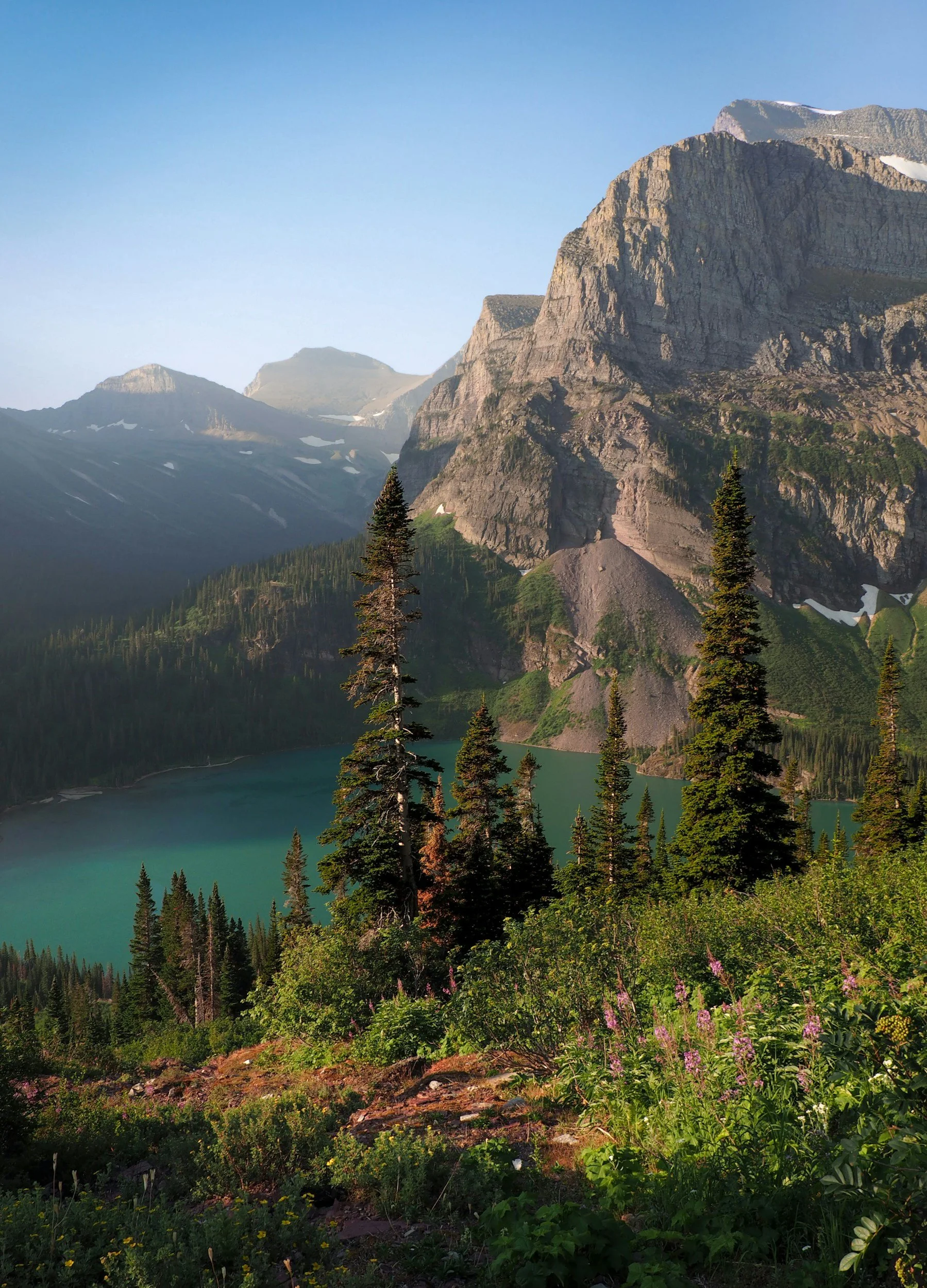 A mountain landscape with tall, rugged peaks, lush green trees, and a turquoise lake in the valley below. The sky is clear and blue.
