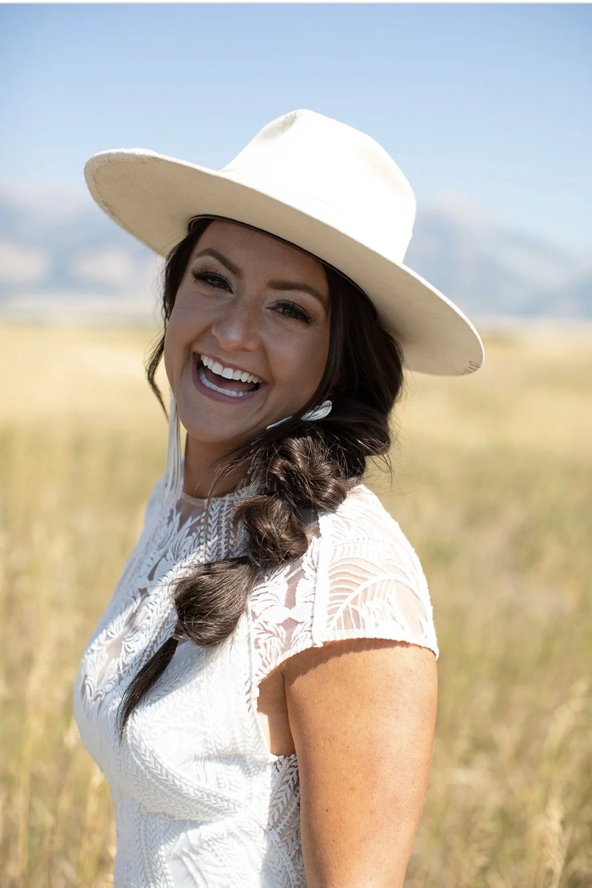 A smiling woman wearing a wide-brimmed hat and a white lace dress standing in a field with a blue sky in the background.