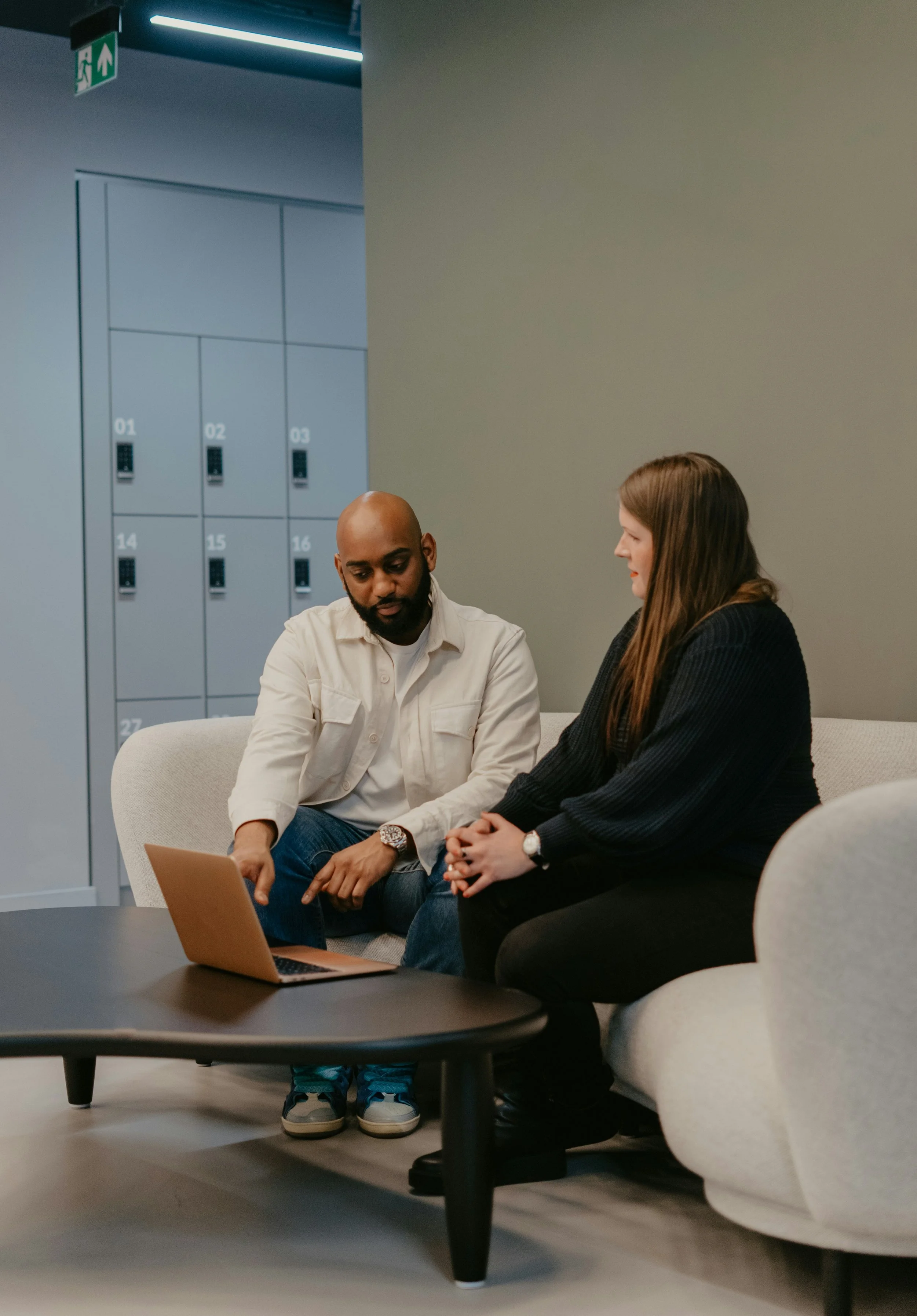 Two people sitting on a couch in a modern workspace or lounge area, looking at a laptop on a small black coffee table in front of them. There are grey lockers with numbered compartments in the background.