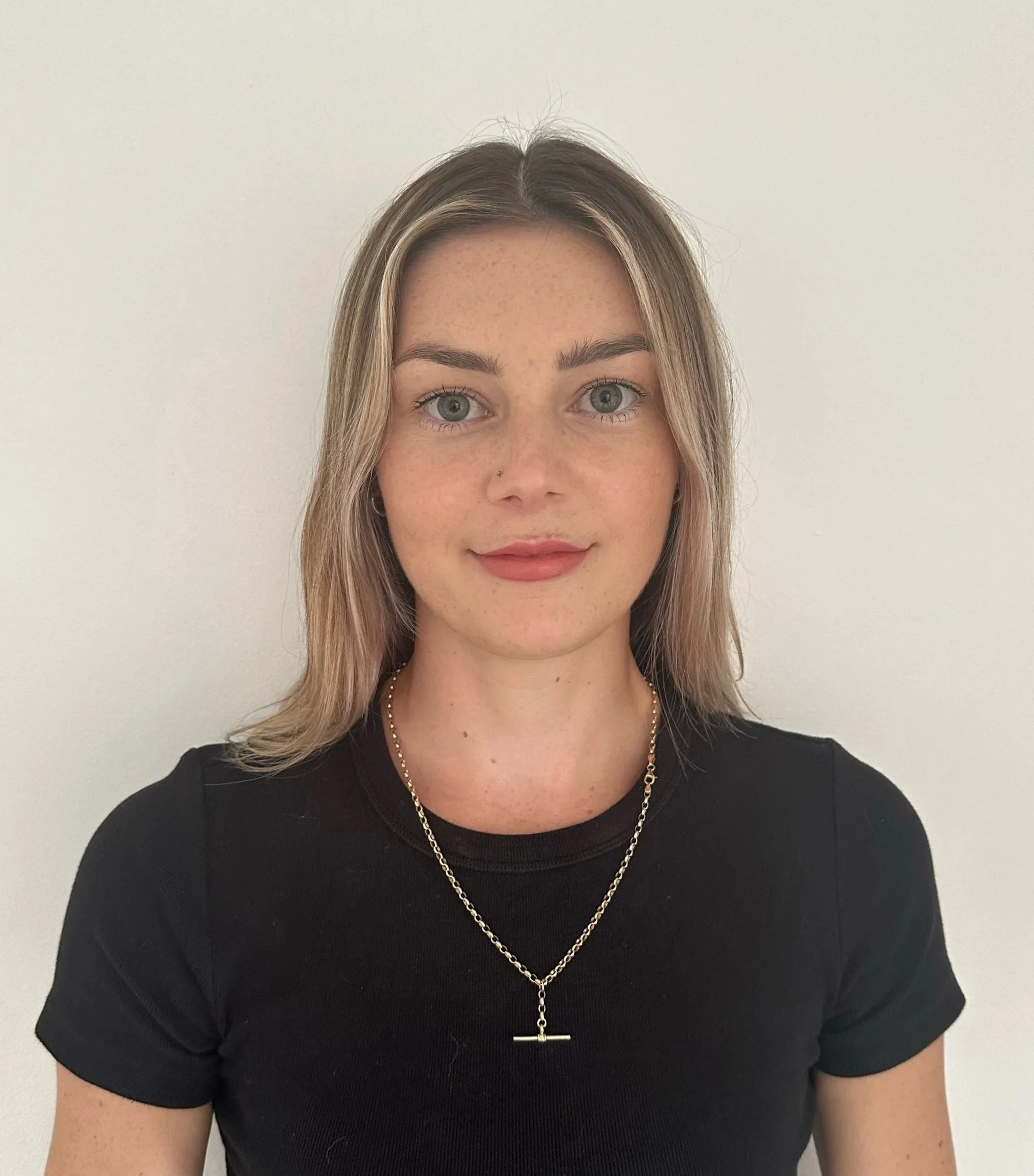 A young woman with light skin, shoulder-length blonde hair, and green eyes, wearing a black t-shirt and a gold necklace with a bar pendant, standing against a plain white wall.