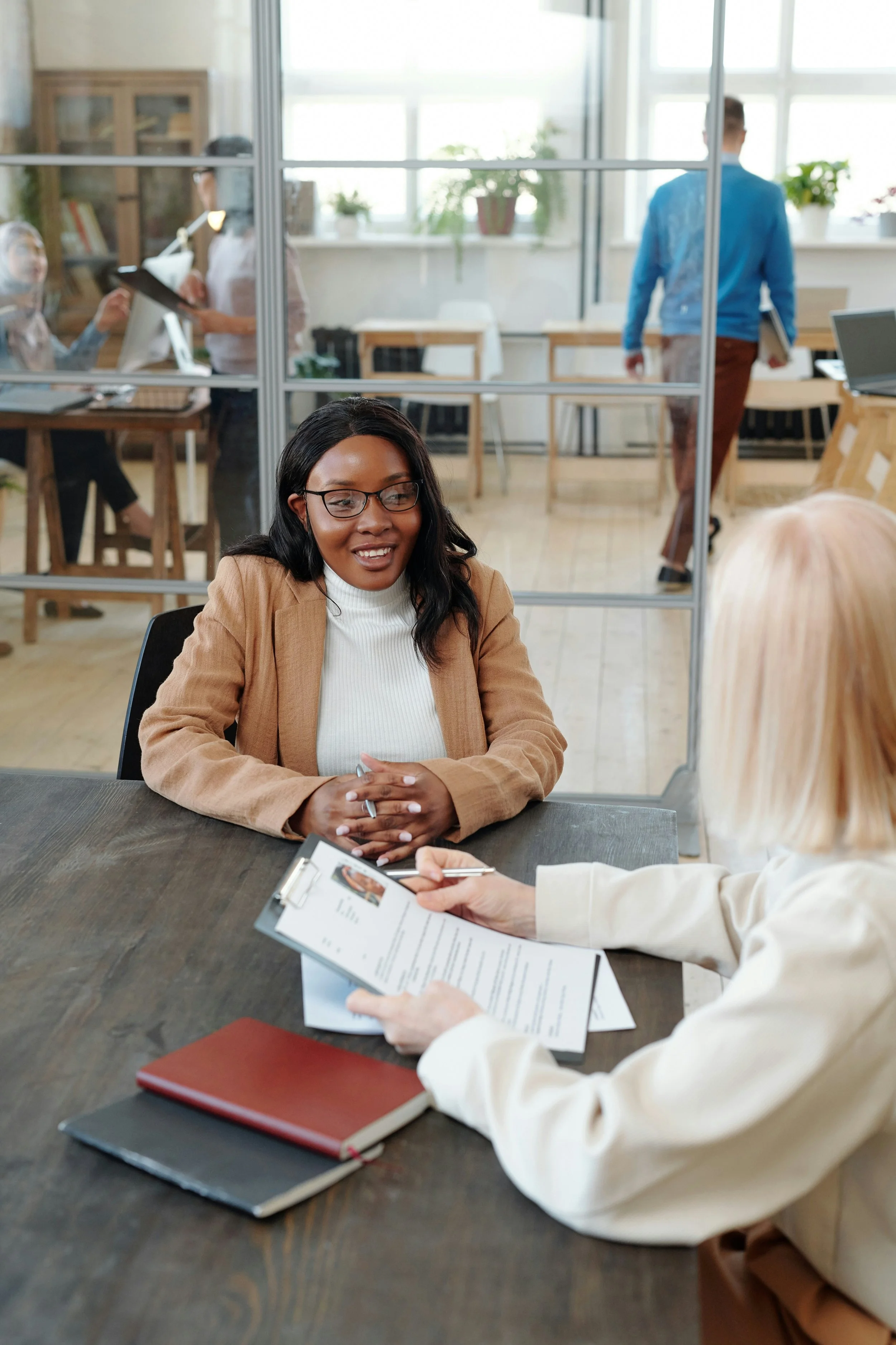 Two women having a meeting in a modern office, one is presenting a resume or document, the other is listening attentively, with books, notebooks, and pens on the table.