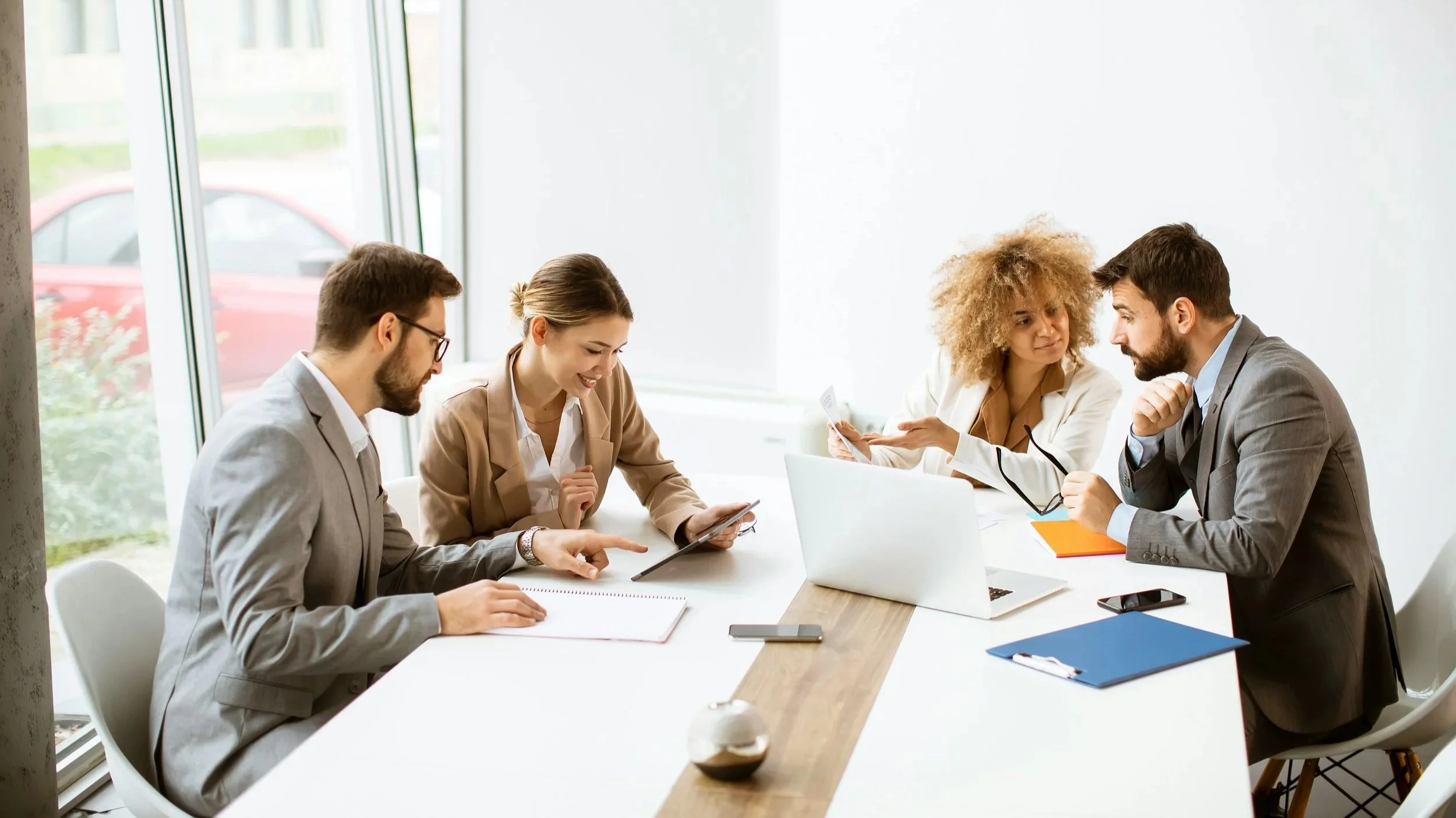 Five professionals in business attire having a discussion at a conference table in a bright room with large windows.