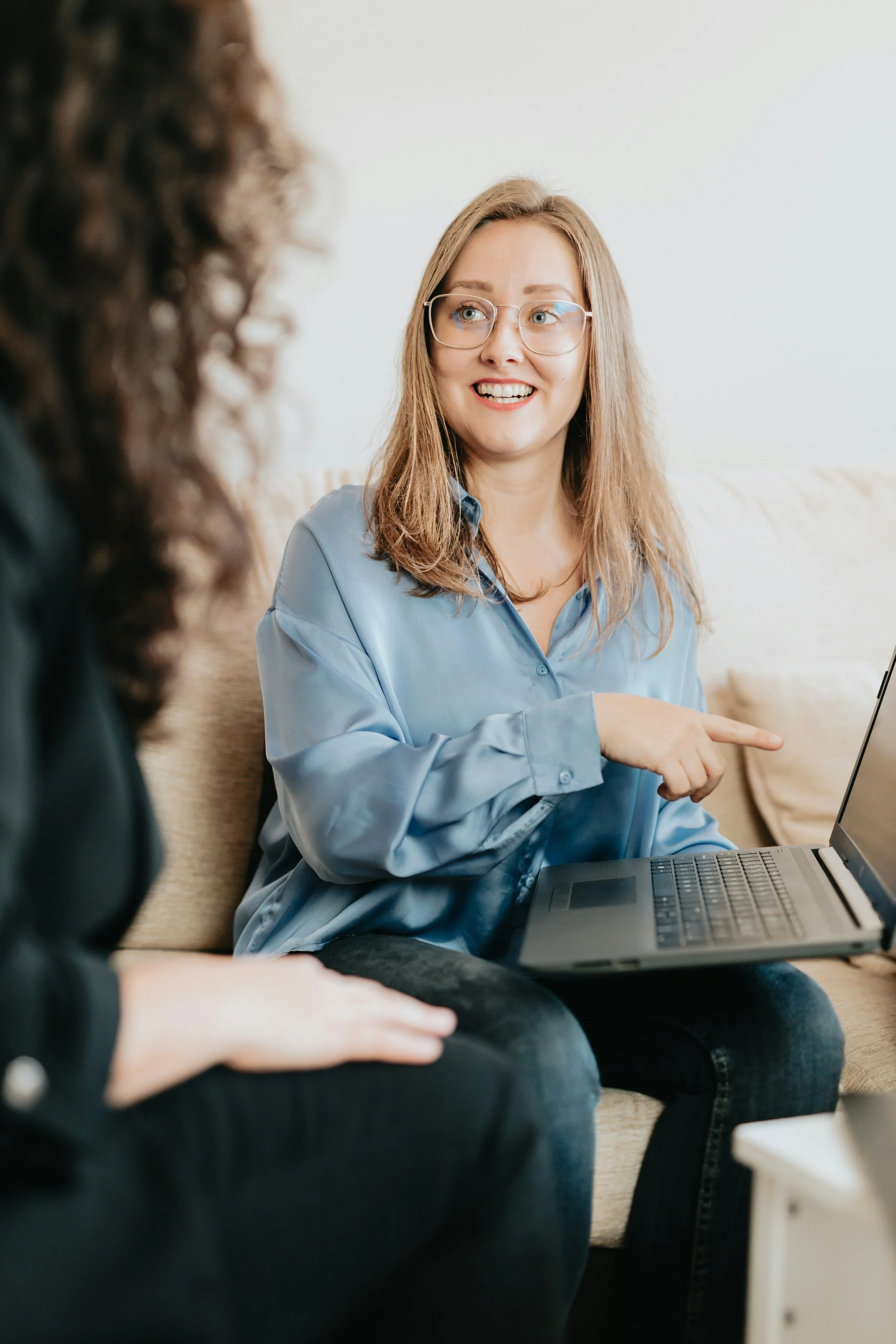 Two women sitting on a beige sofa, having a discussion. The woman on the right is wearing glasses, a blue silk blouse, and is pointing at a laptop. The woman on the left has curly hair and is dressed in black.