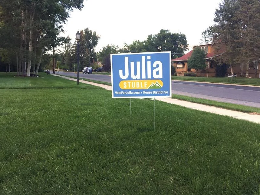 A campaign sign on a grassy lawn supporting Julia Stuble for House District 54, with trees, a sidewalk, and houses in the background.