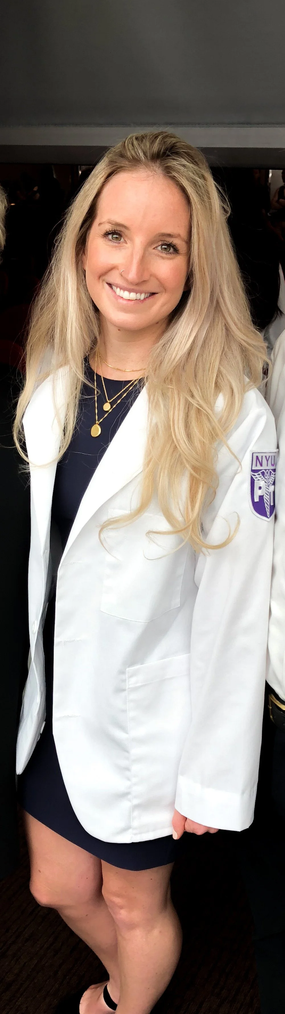 Young woman with long blonde hair, wearing a white NYU medical coat with a purple patch, a black dress, and layered gold necklaces, smiling indoors.