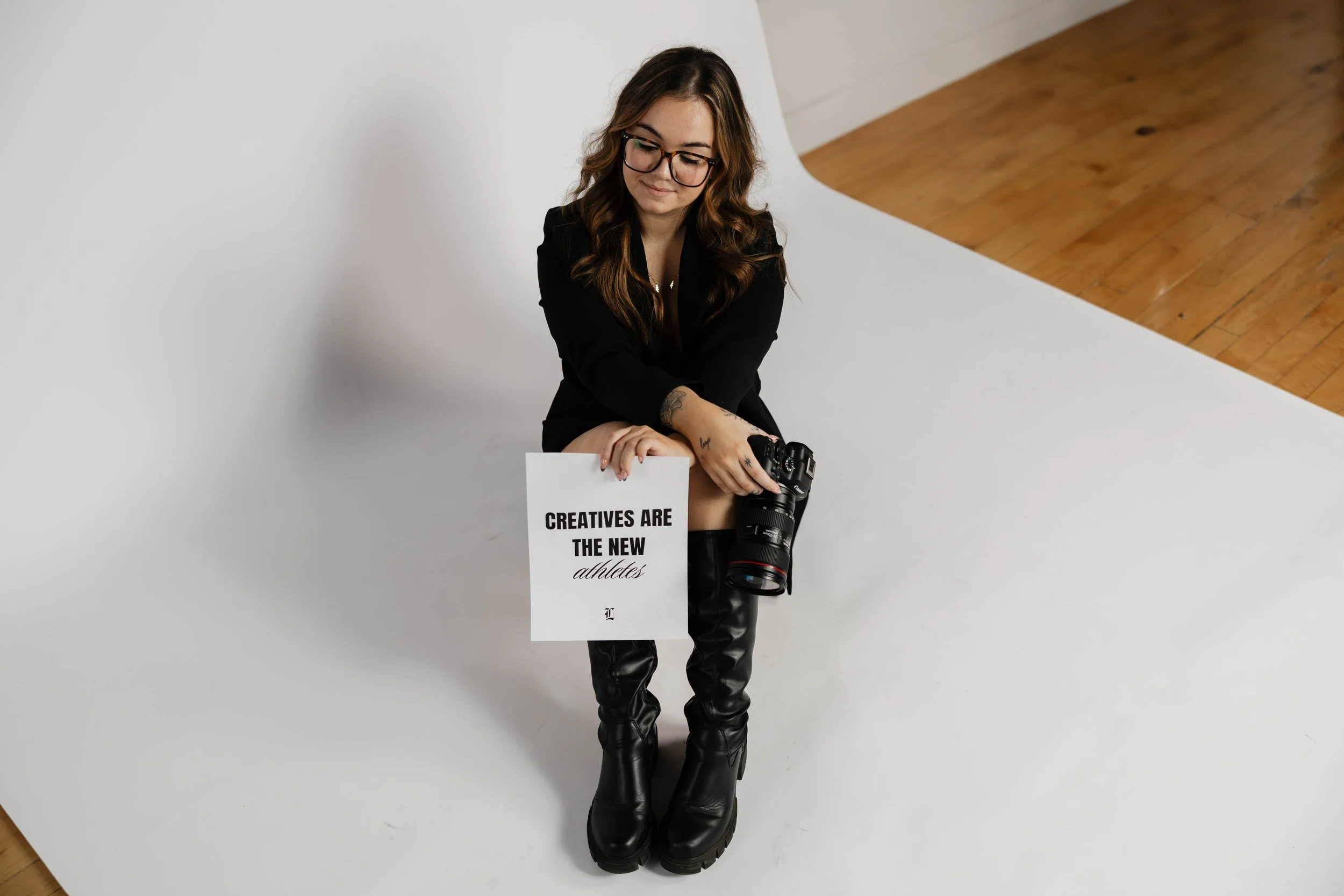 A woman seated on a white backdrop wearing black boots and holding a camera and sign that reads 'Creatives Are The New Athletes'.