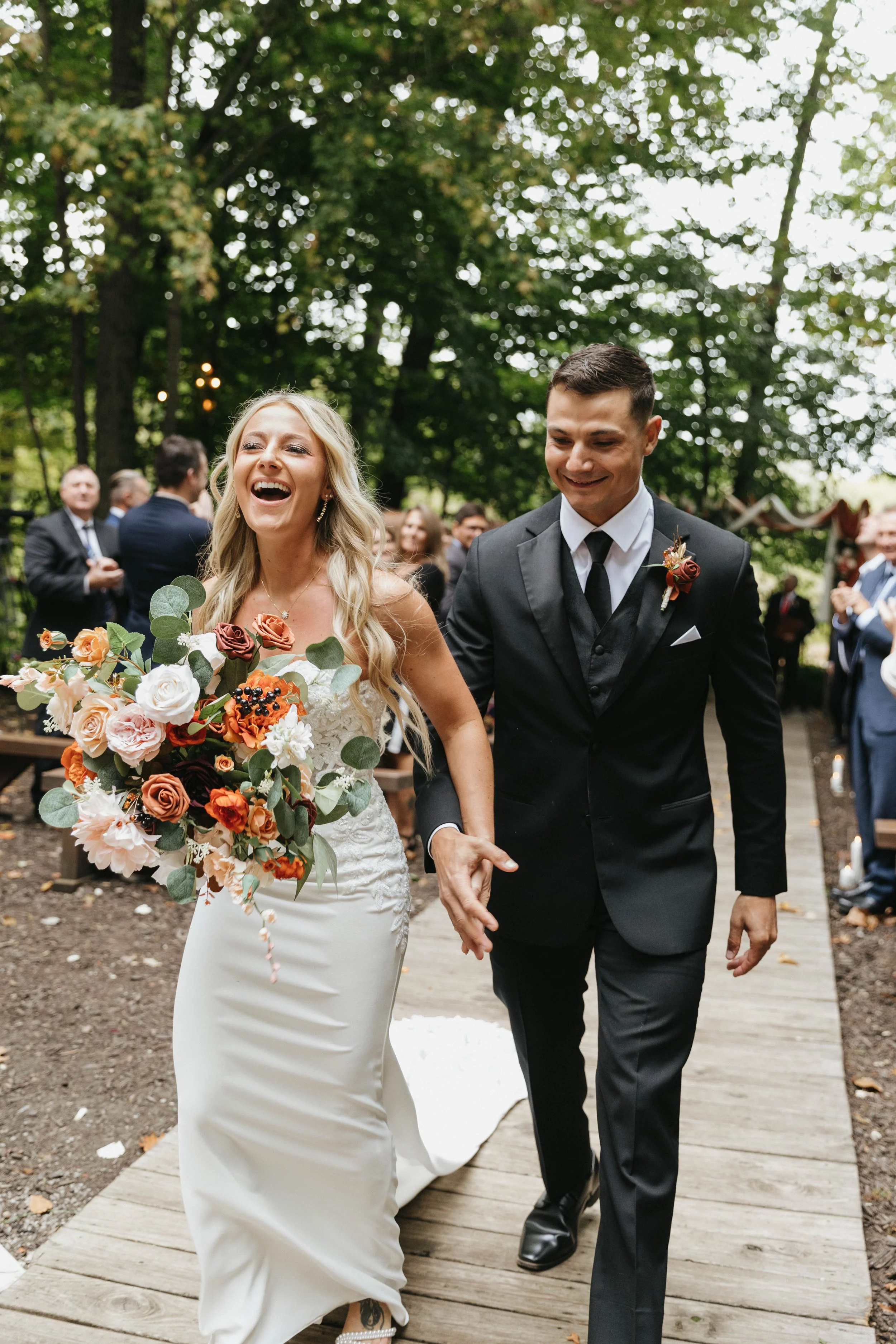 A newlywed couple walking down the aisle outdoors; the bride is holding a large bouquet of flowers and laughing, while the groom next to her is smiling. Guests are clapping in the background amid green trees.
