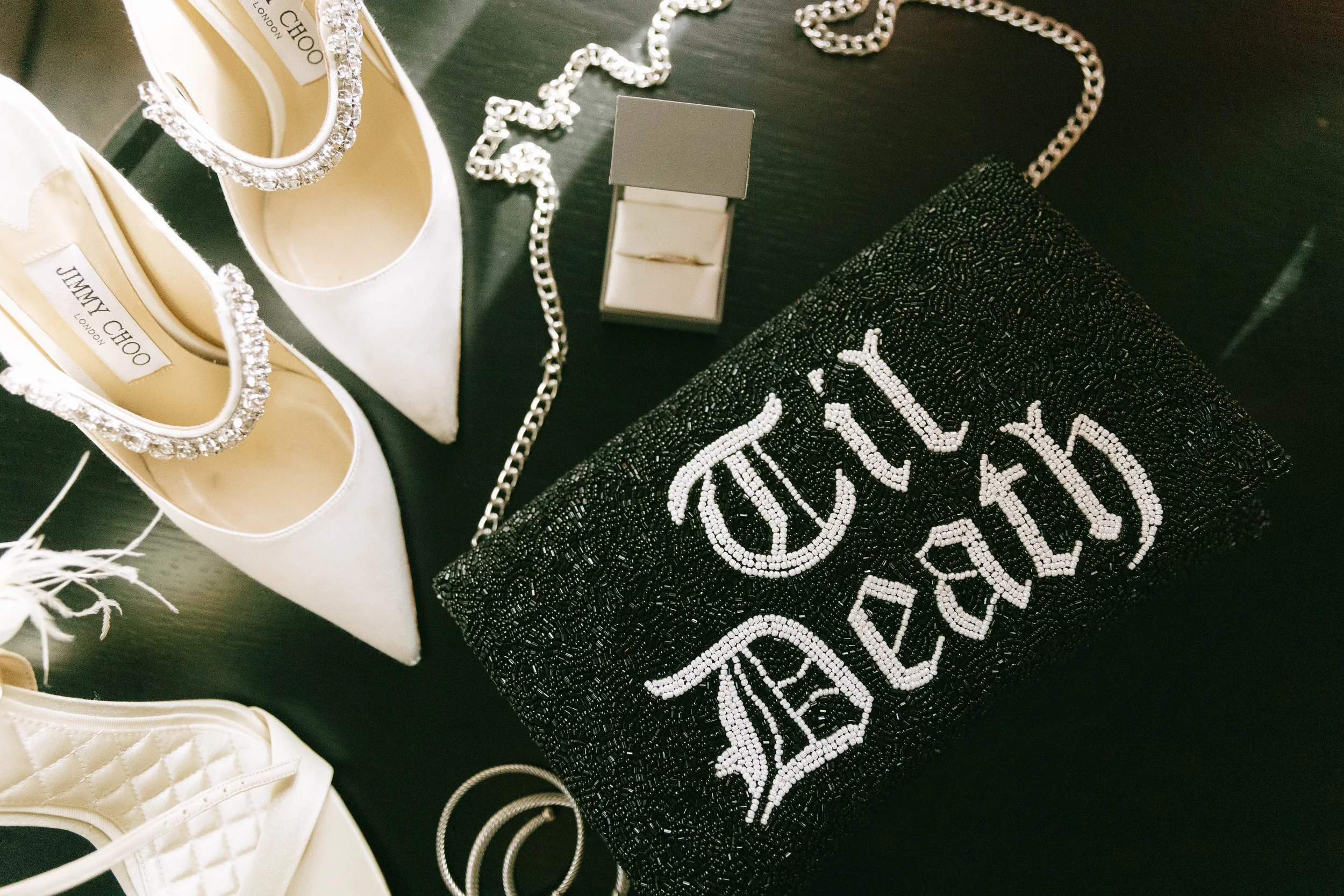 A collection of luxury accessories on a black surface, including white pointed toe shoes with rhinestone ankle straps, a black beaded clutch with white embroidery, a small jewelry box with rings inside, and a silver chain necklace.