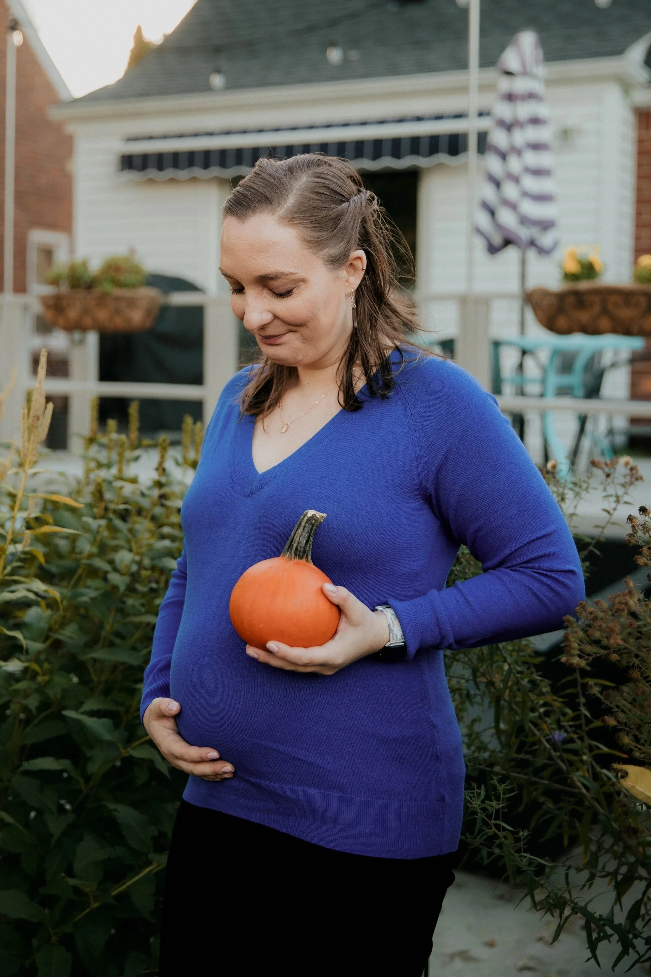 A pregnant woman in a blue sweater holding a small pumpkin in her hand standing in a garden.
