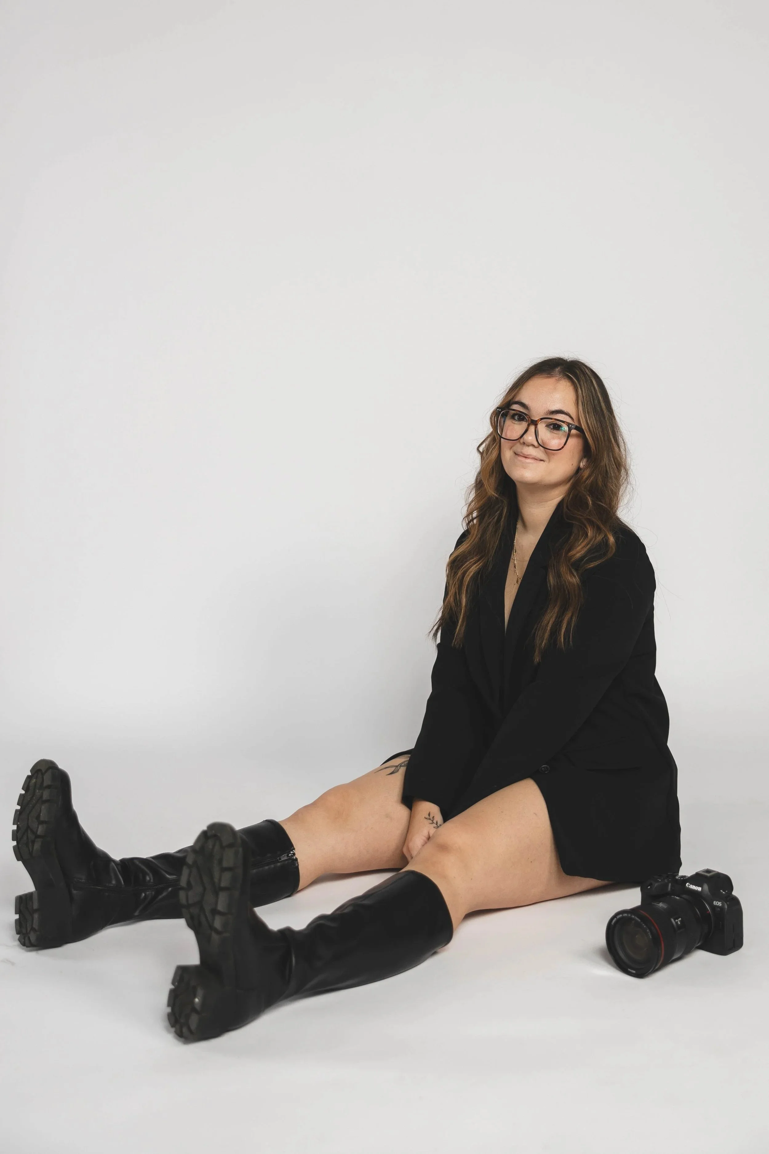 Woman sitting on the floor wearing a black blazer and boots, with a camera next to her, against a plain white background.