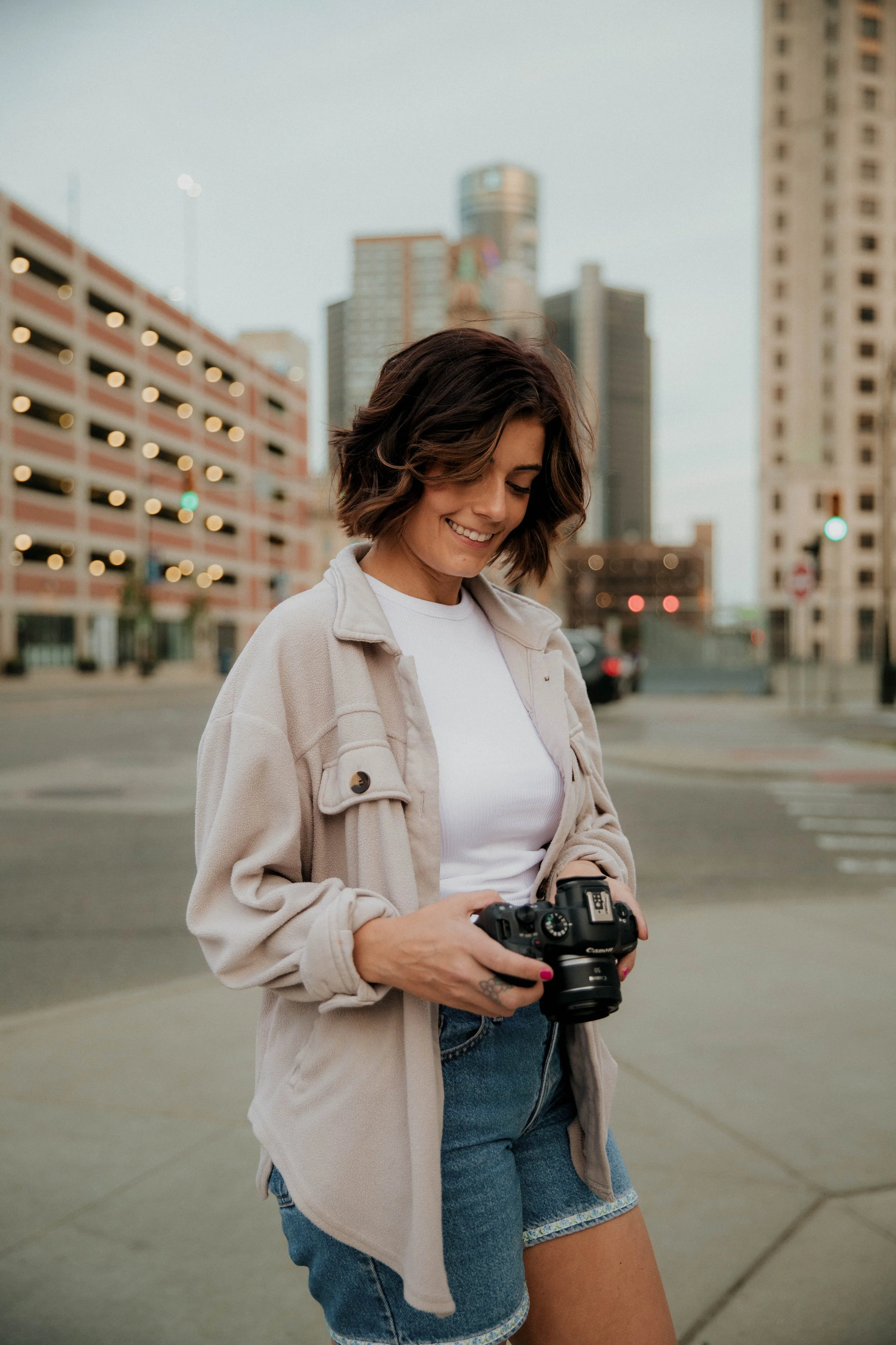 A smiling woman with short brown hair stands on a city sidewalk holding a DSLR camera, wearing a white shirt, beige jacket, and denim shorts, with tall buildings in the background.