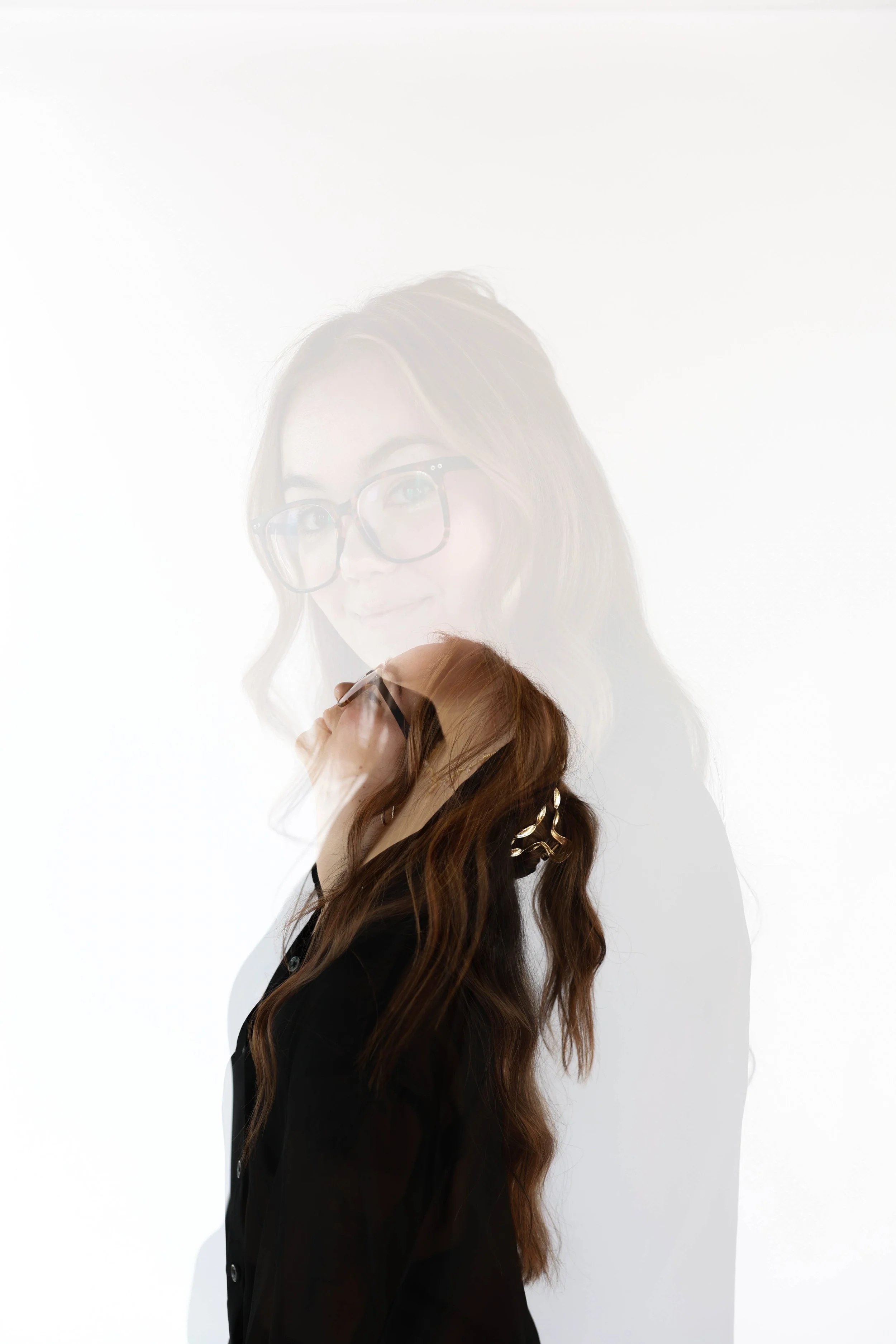 A woman with long wavy hair wearing glasses and a black shirt, superimposed in multiple exposure, creating a layered effect against a white background.