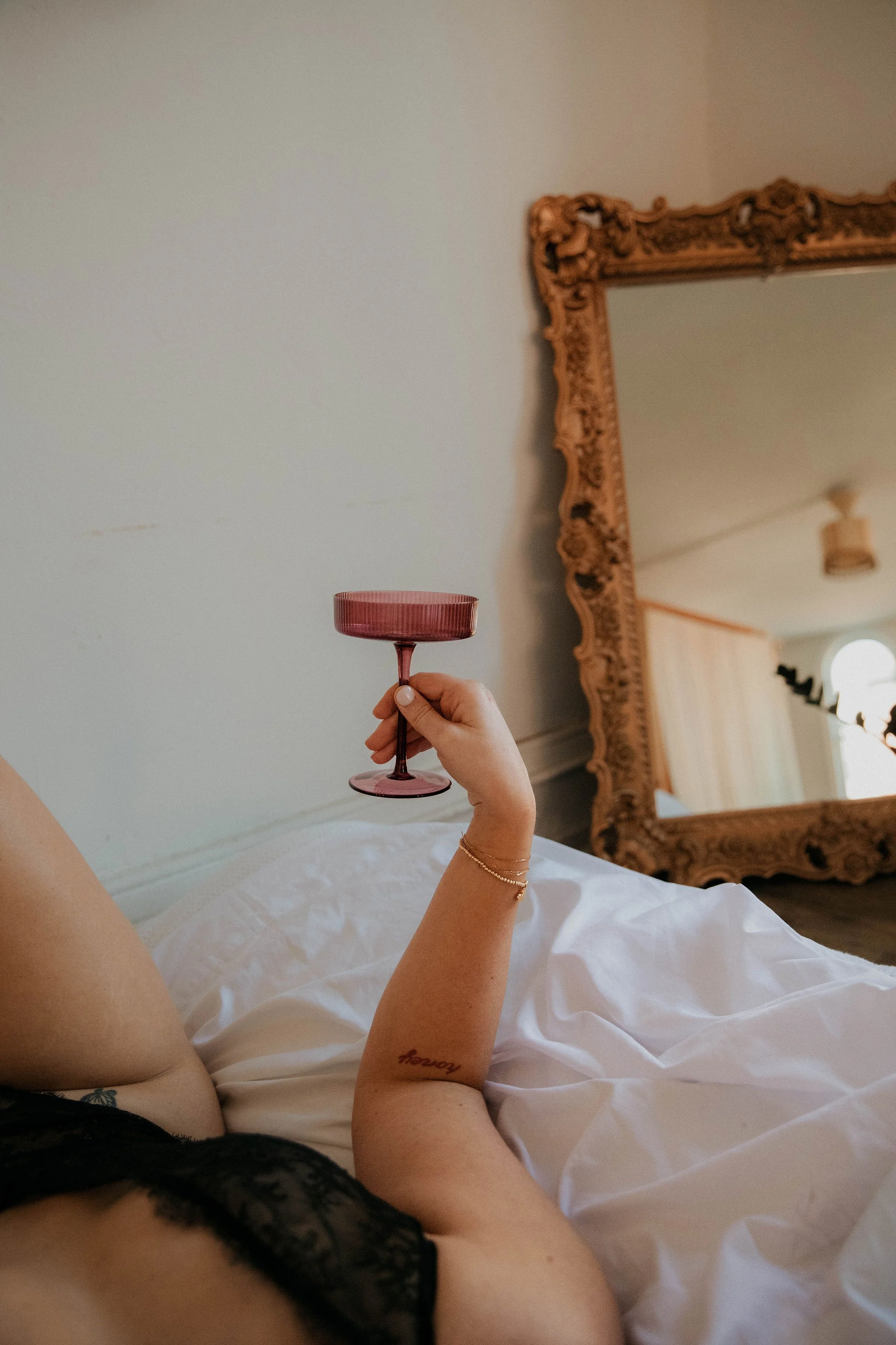 Person lying on bed holding up a pink chalice in a bedroom with a large ornate mirror.