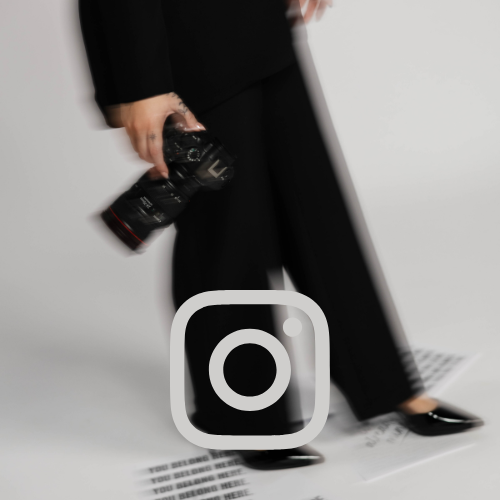 Person in black attire holding a professional camera while standing on a white surface with a blurred plain background.