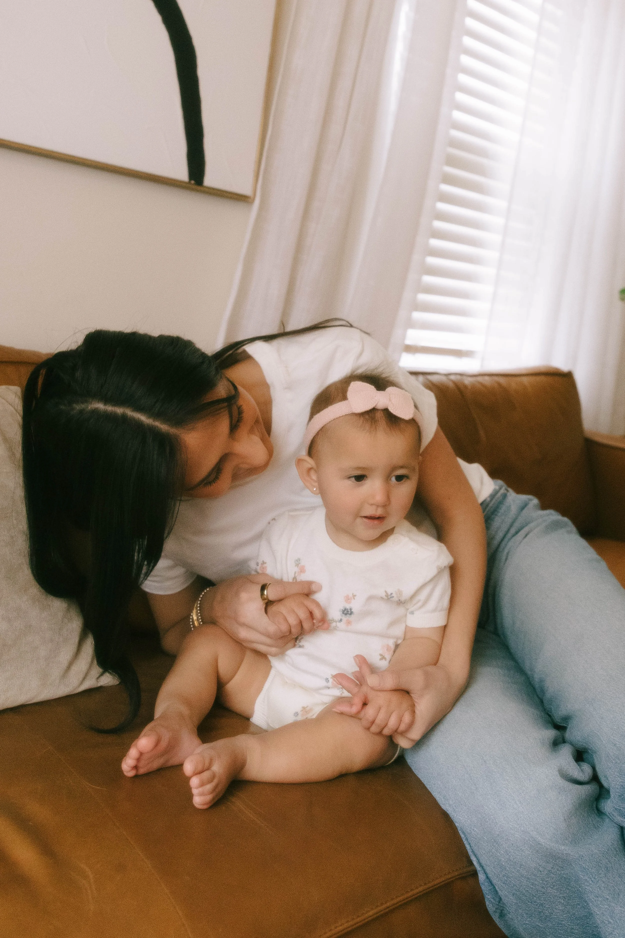 A woman and a young girl are sitting on a brown couch, with the woman leaning over and holding the girl. The girl is wearing a pink headband and a white shirt with floral embroidery. The background shows white curtains and a picture frame on the wall.