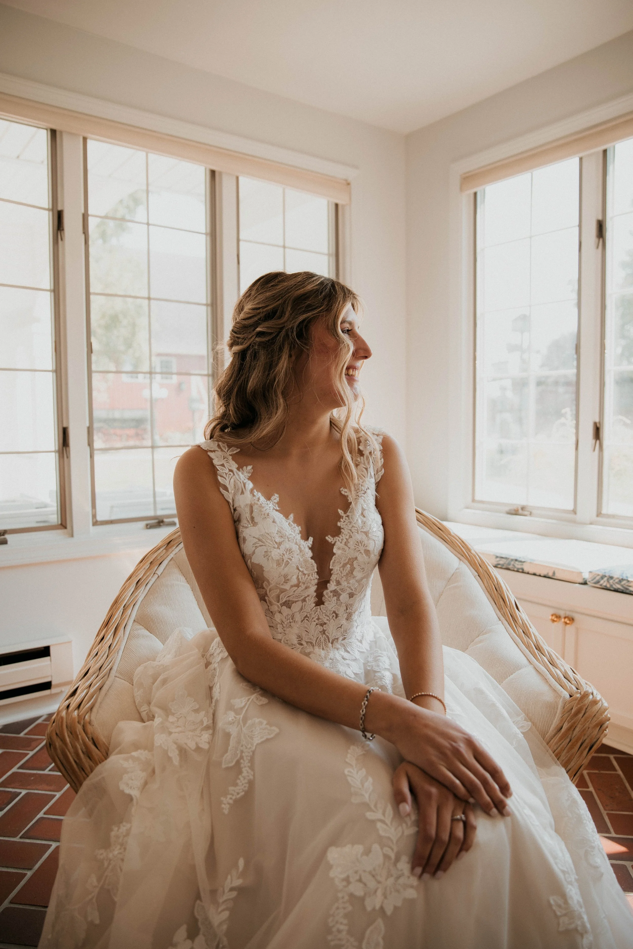 A bride in a lace wedding gown sitting on a wicker chair, smiling and looking to the side, inside a bright room with large windows.
