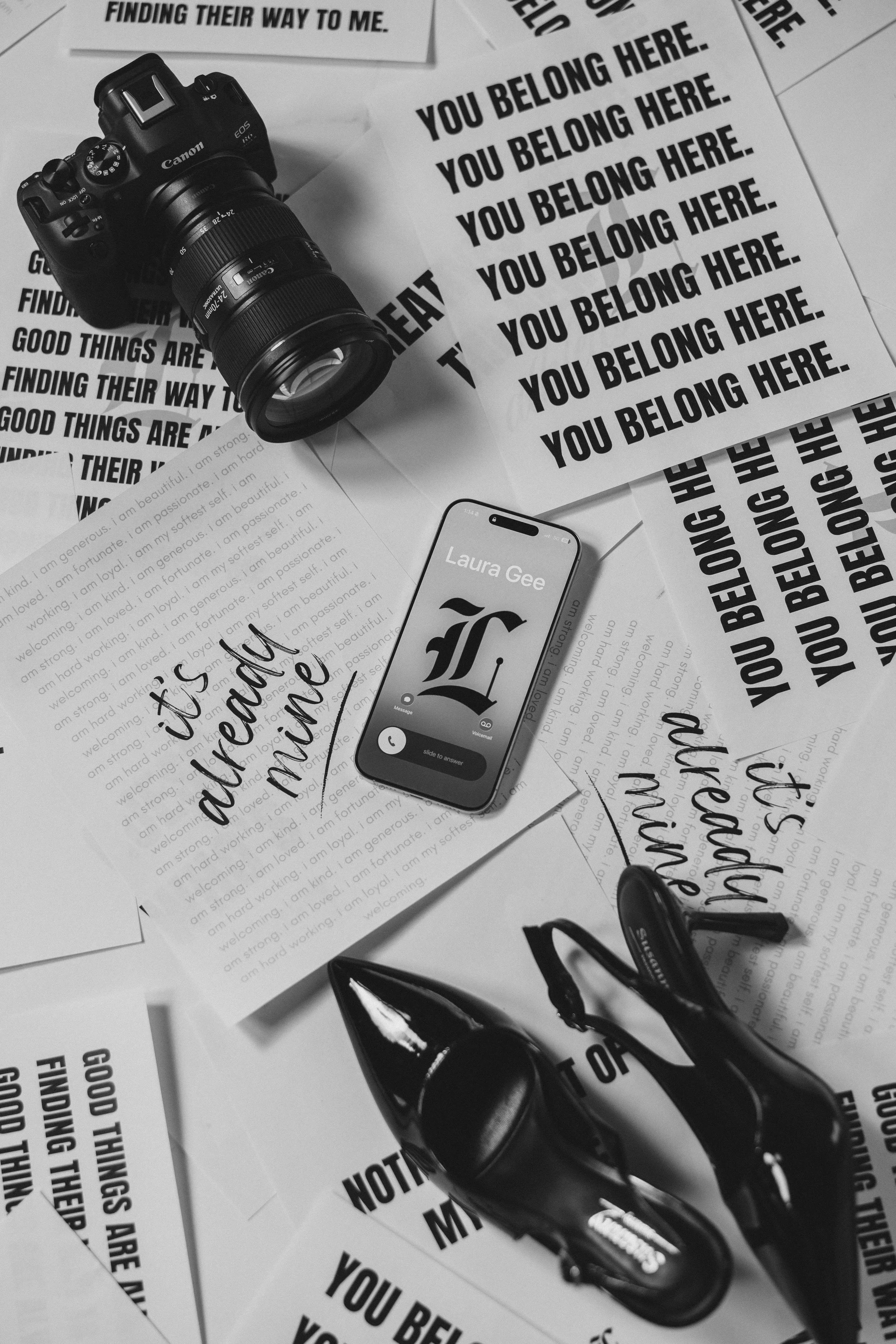 Black and white photo of a cluttered desk with a camera, a high heel shoe, a smartphone displaying a call from Laura Gee, and multiple sheets of paper with bold printed phrases like 'You belong here' and handwritten notes.