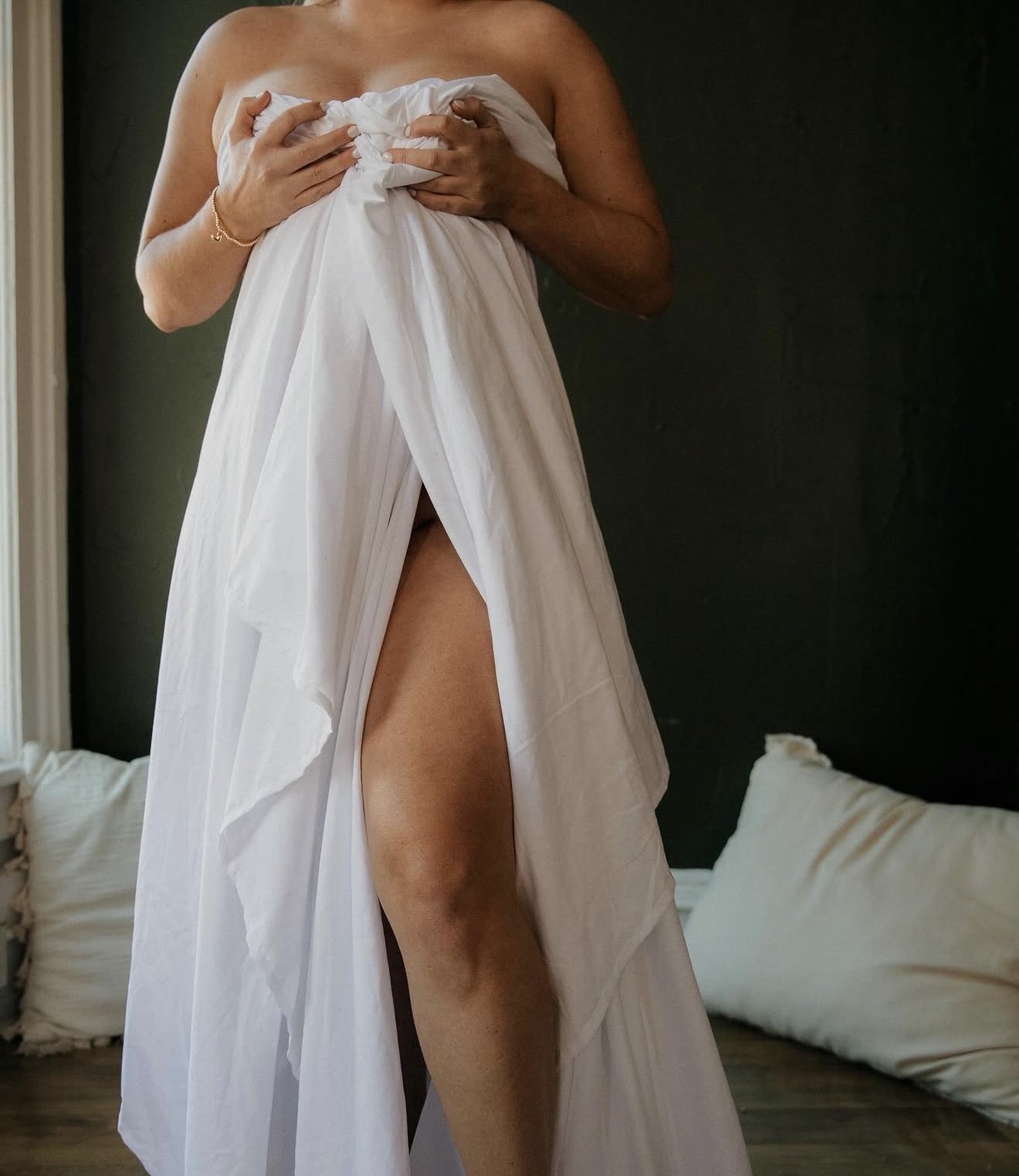 A woman holding a white sheet, partially covering her body, revealing her legs and midsection, standing indoors against a dark wall.