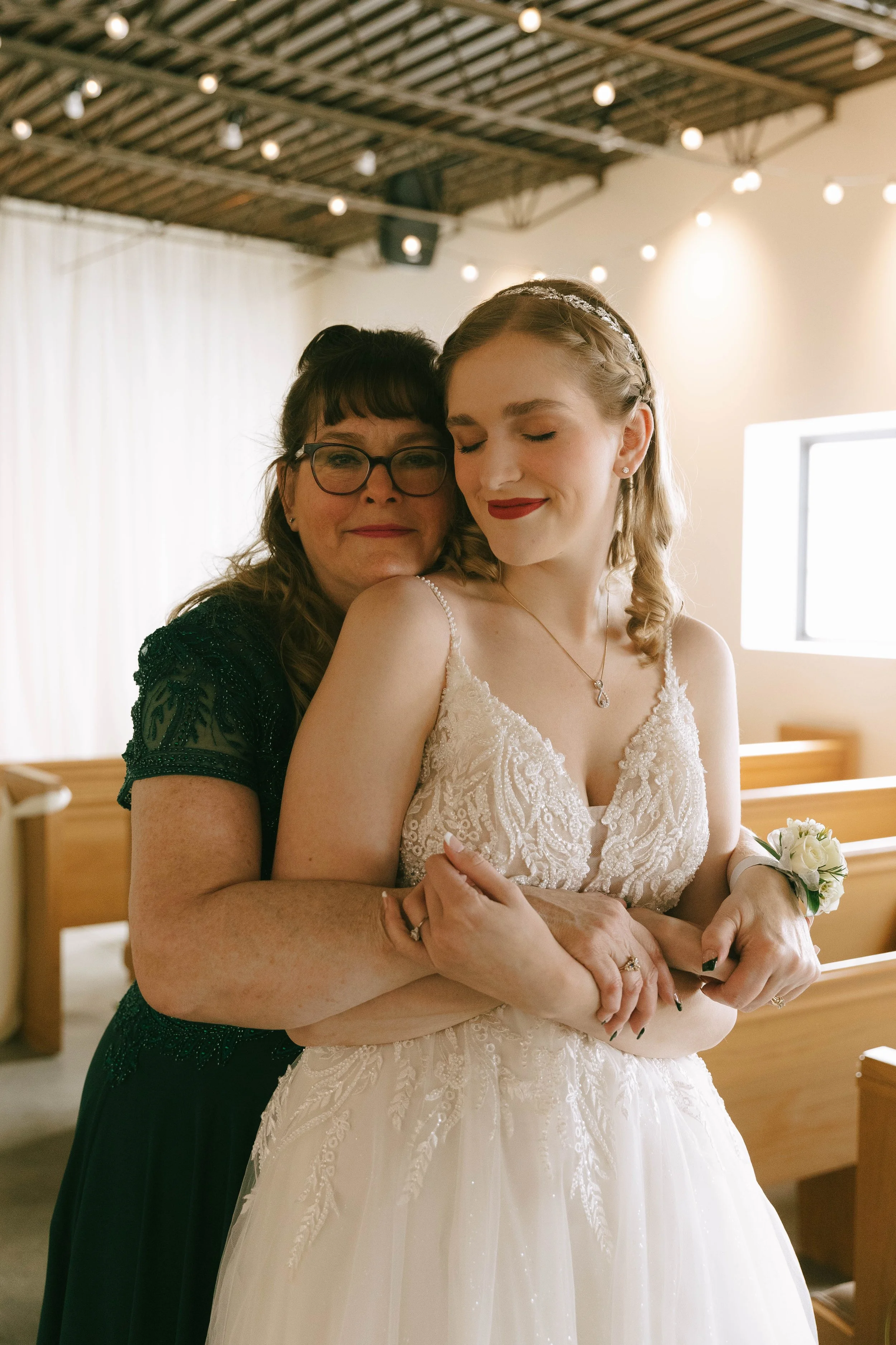 A bride in a white wedding gown and a woman hugging her from behind inside a church with wooden pews and string lights.