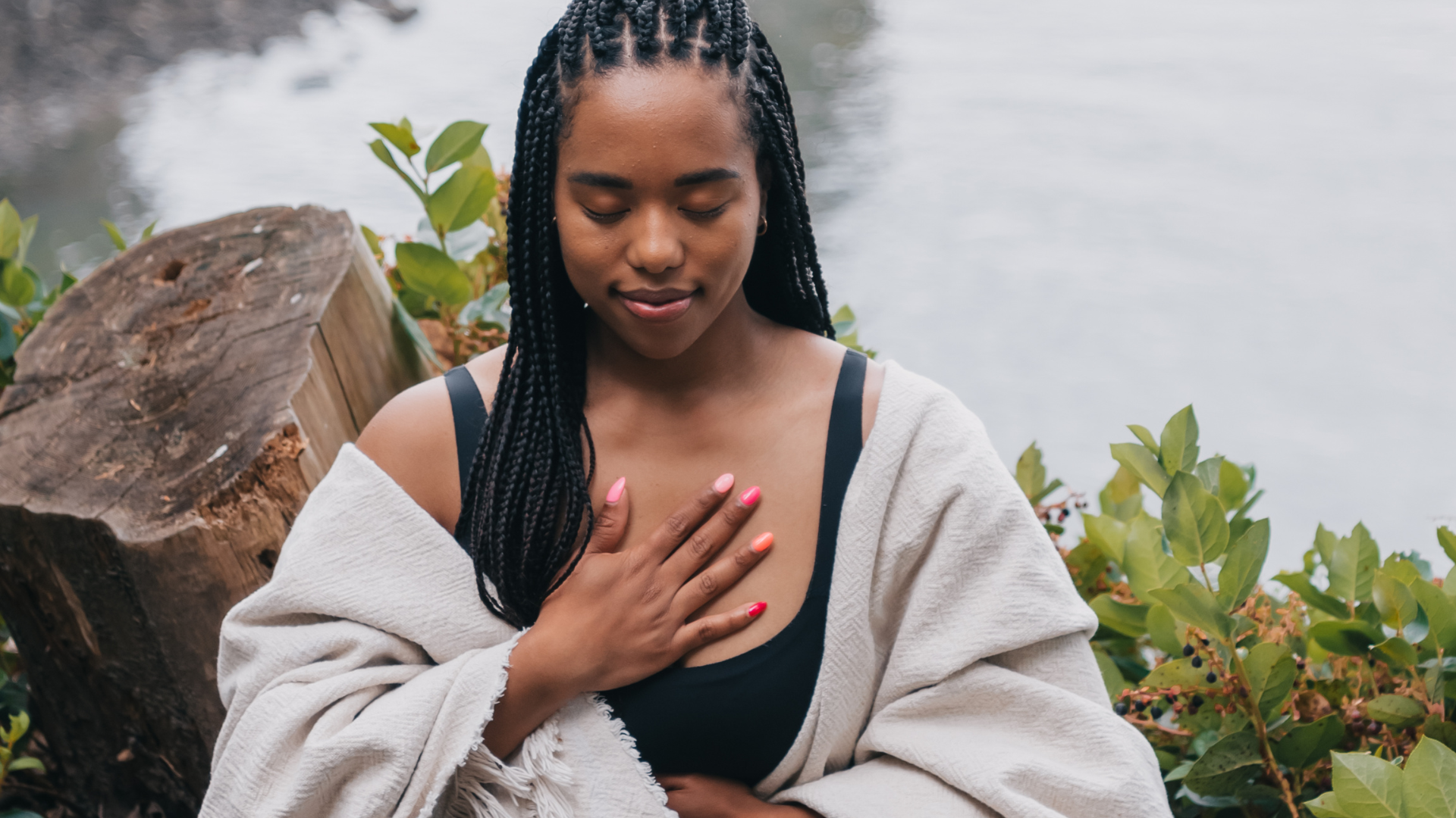 A young woman sits by a lake and practices breathwork for anxiety, laying her hands on her chest.