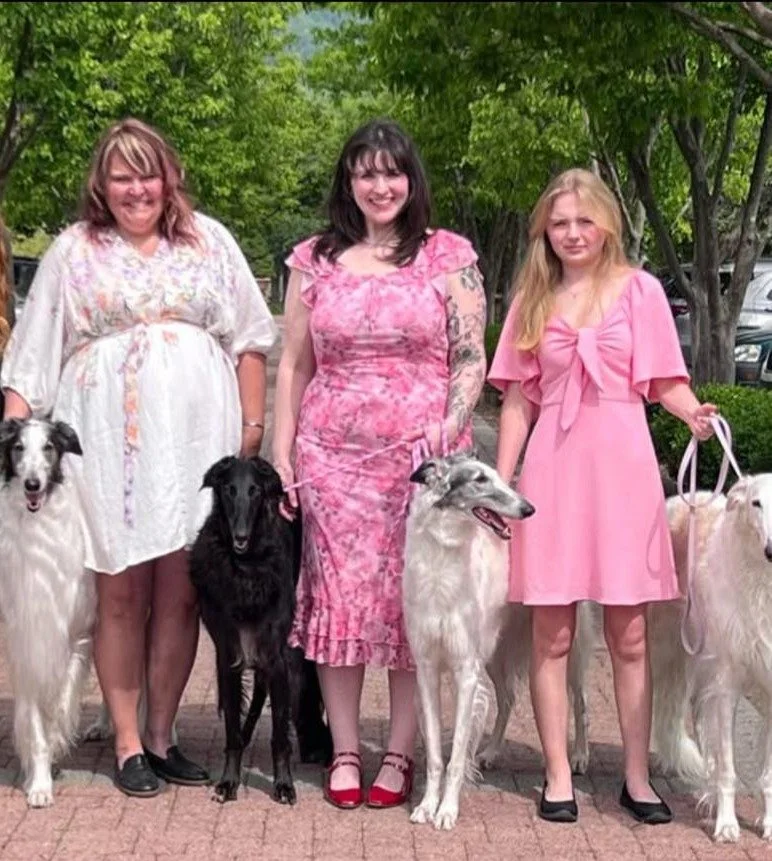 Three women standing outdoors with four dogs on leashes, in front of green trees and parked cars.
