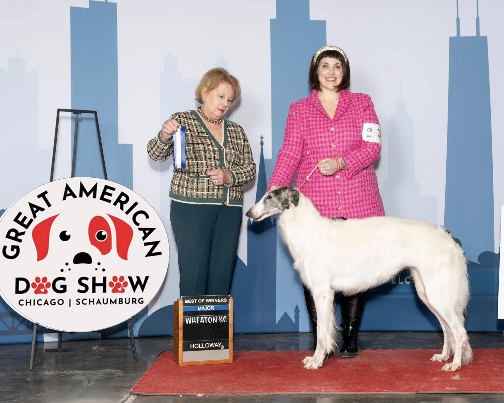 A woman in a pink blazer holding a dog leash with a Borzoi dog, standing next to another woman in a plaid jacket holding a ribbon, at the Great American Dog Show in Chicago Schaumburg with a city skyline backdrop.