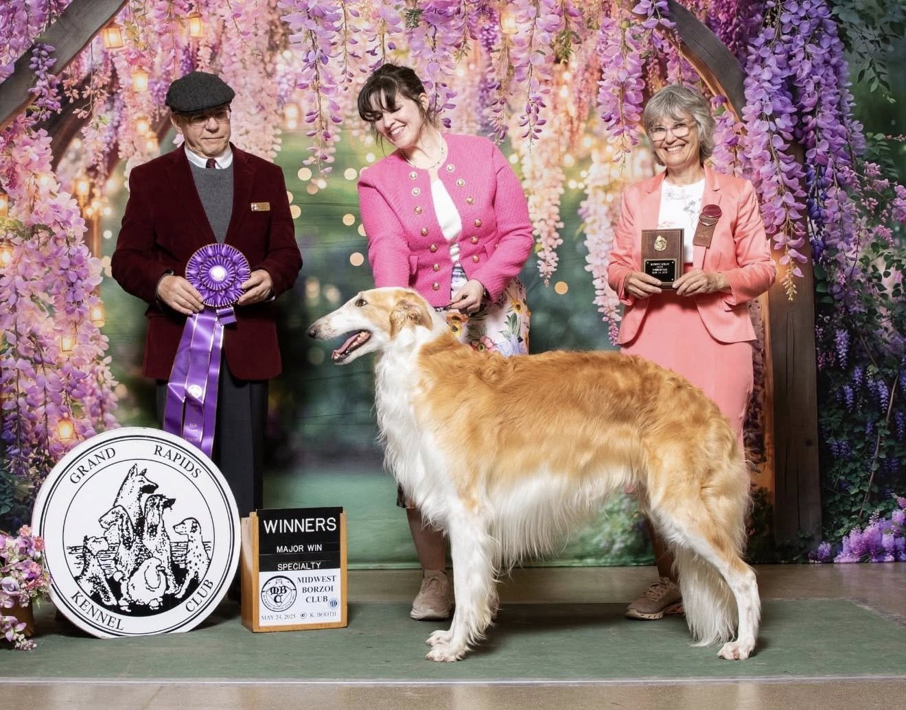 Borzoi standing in front of three women during an awards presentation at the Grand Rapids Kennel Club dog show, with ribbons, trophies, and floral backdrop visible.