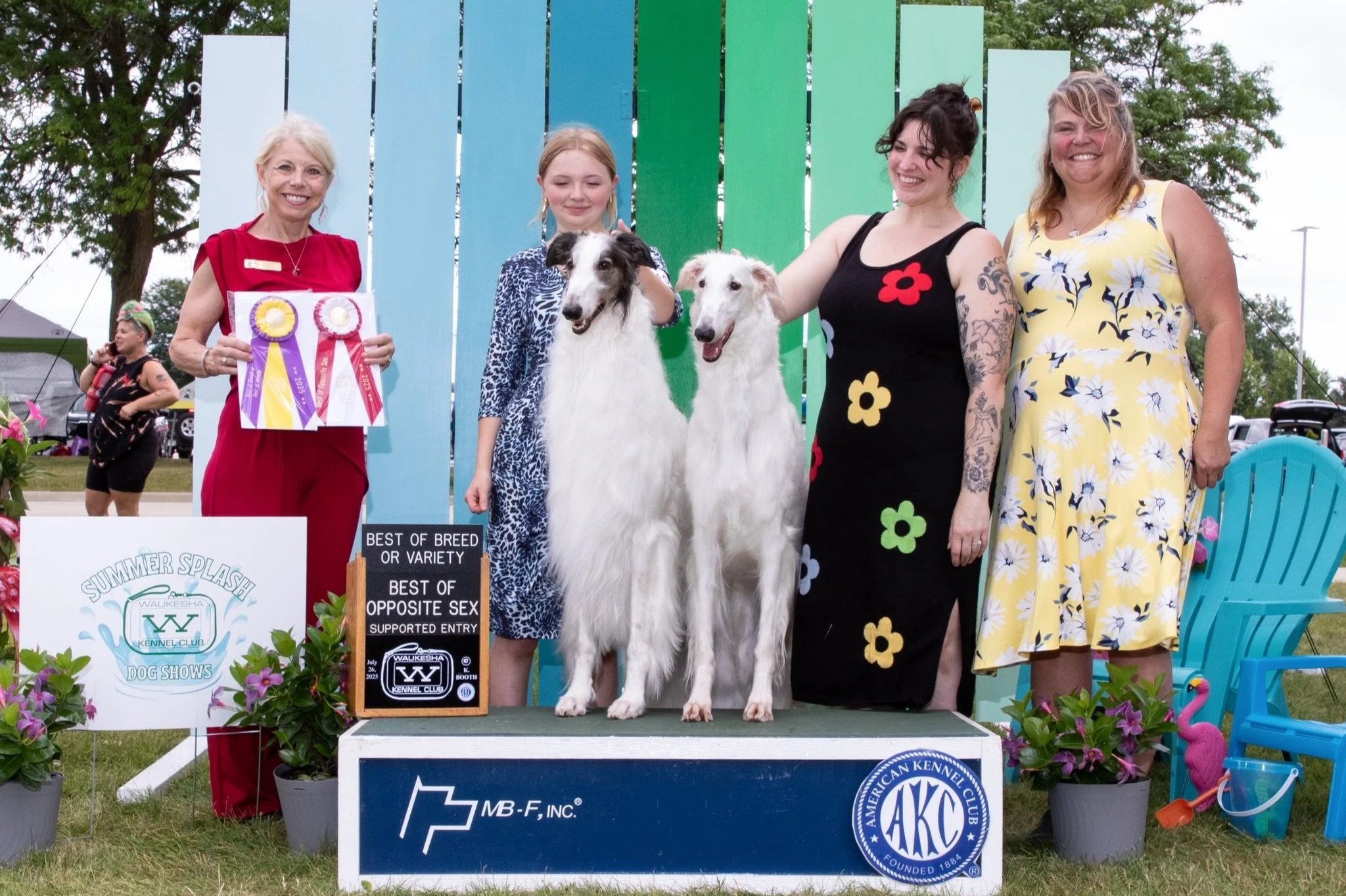 Four women posing with two Borzoi at an outdoor dog show awards presentation, with Best of Breed ribbons and signage visible, set among trees and show tents.