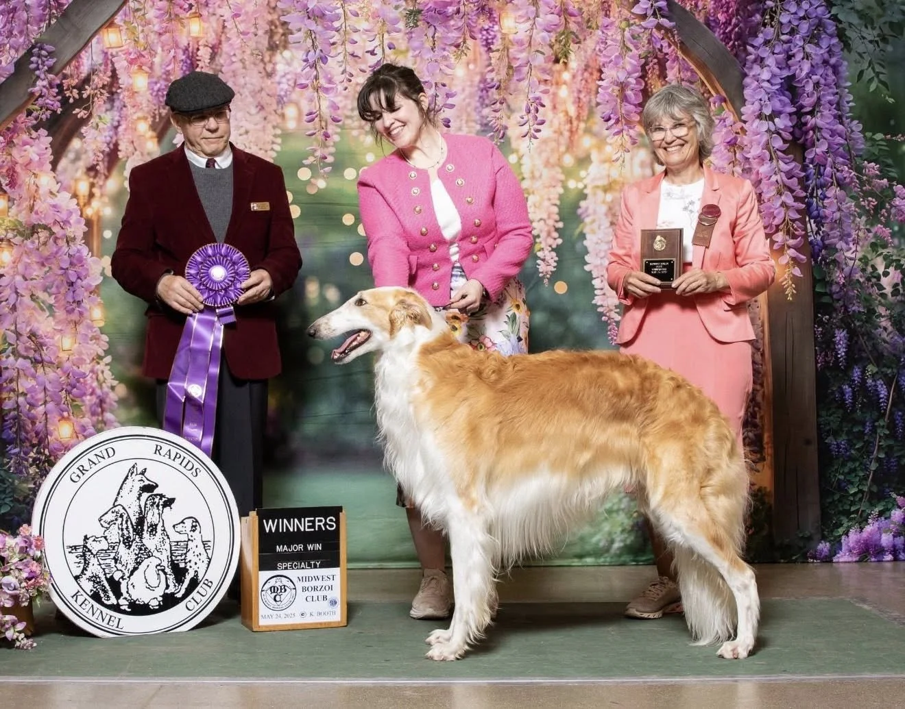 A group of three women and a Borzoi dog standing on a green platform at a dog show. The women are dressed in formal attire, holding awards, with a colorful floral and fairy light backdrop. They are smiling and celebrating the dog's win, with a sign that reads 'Winners' and a large circular emblem of the Grand Rapids Kennel Club. The woman in the middle is holding the dog, which is standing proudly.