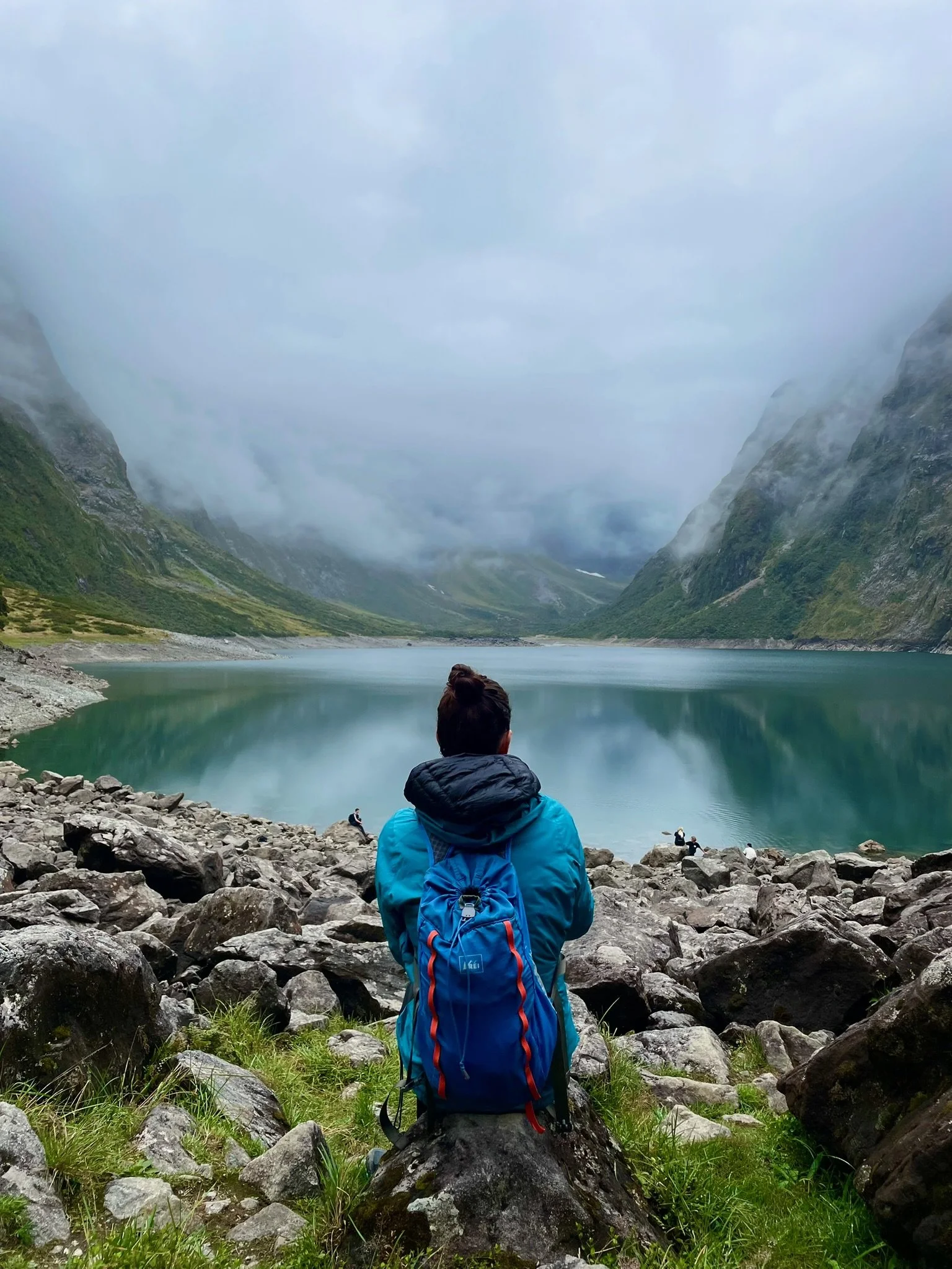 A person with a blue backpack sitting on rocks in front of a calm lake surrounded by green mountains and cloudy sky.