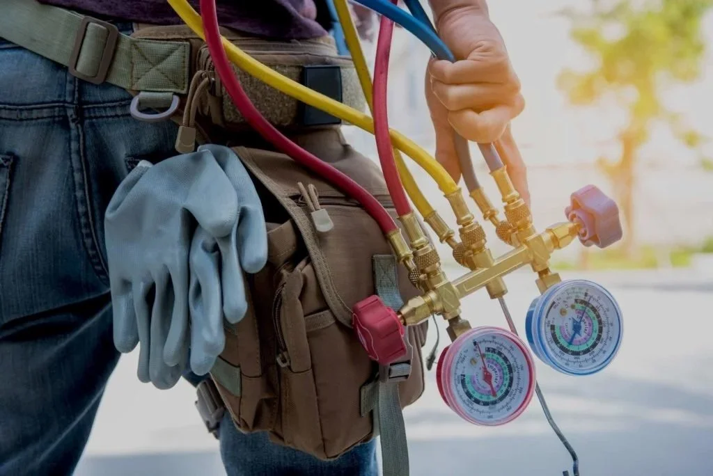 A worker in jeans standing outdoors, holding a manifold gauge set with red, yellow, and blue hoses, used for HVAC or refrigeration systems. The worker has gloves attached to a tool belt.