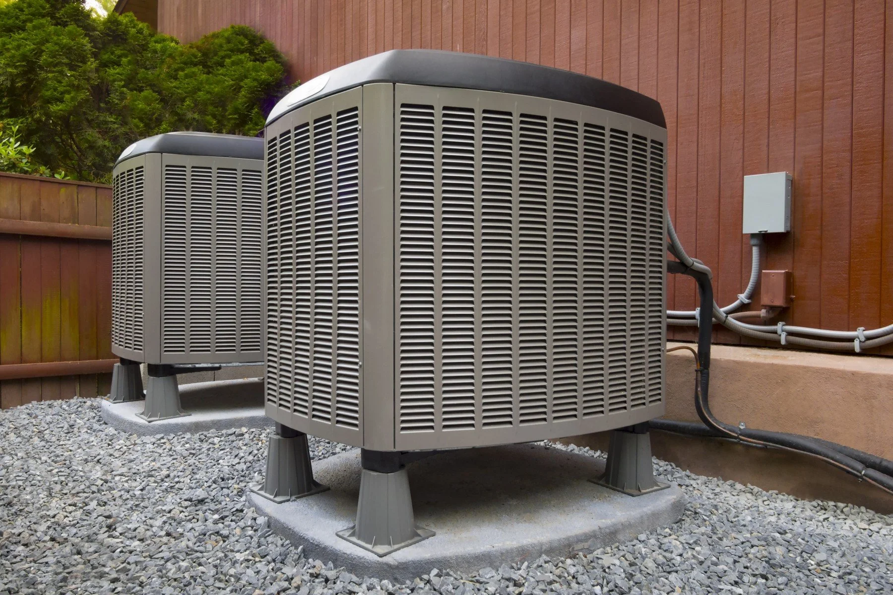 Two outdoor HVAC units installed on a gravel surface against a wooden fence with trees in the background.