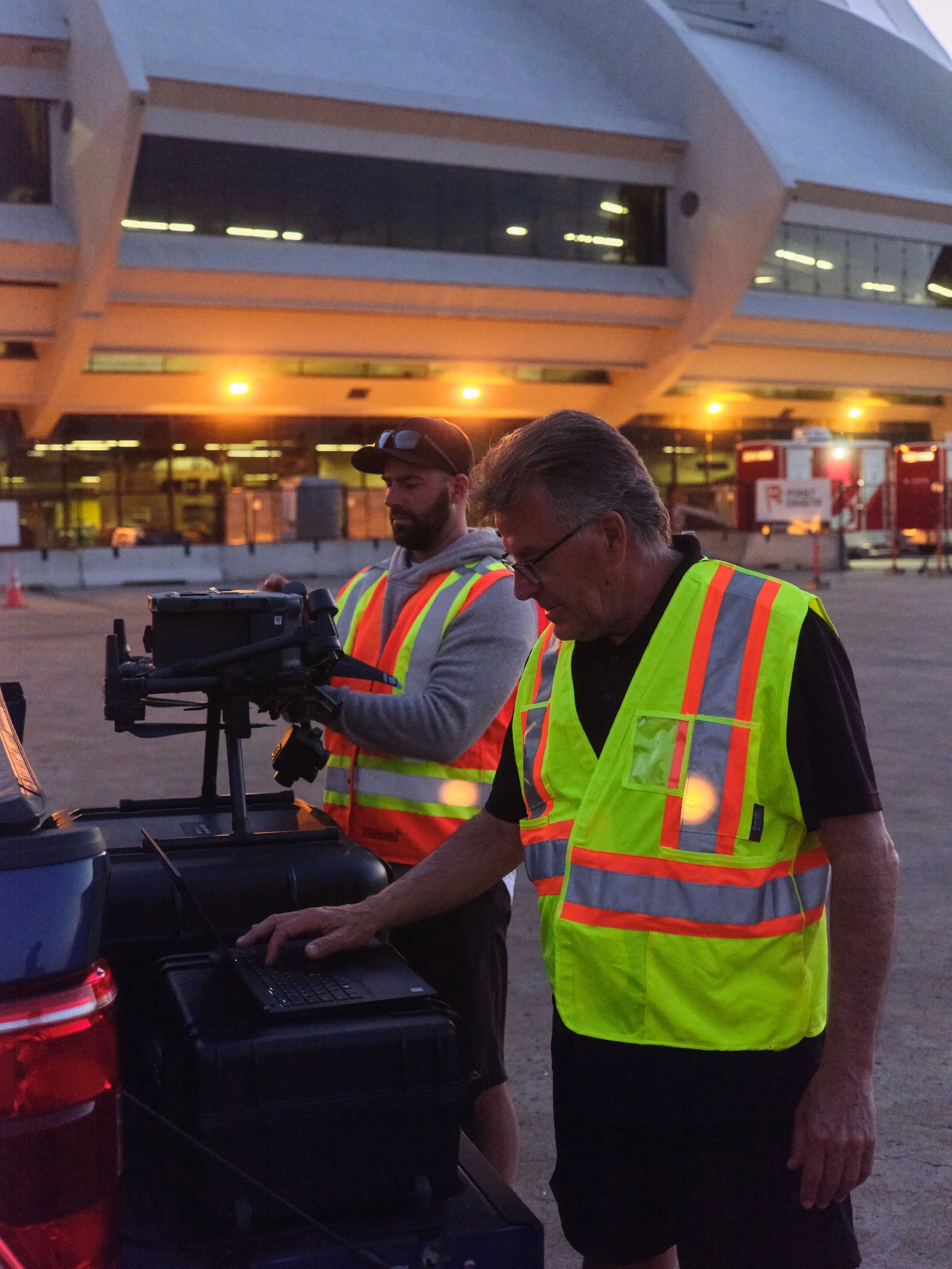 Two men in reflective safety vests working on equipment near an open trunk of a car outside during evening or early morning, with a modern building in the background.