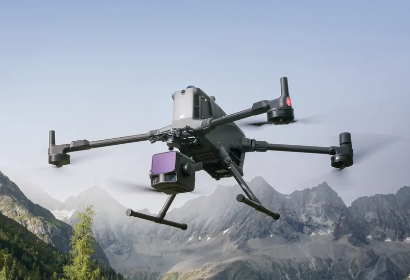 A quadcopter drone flying in the sky over mountainous terrain with trees and rocks.