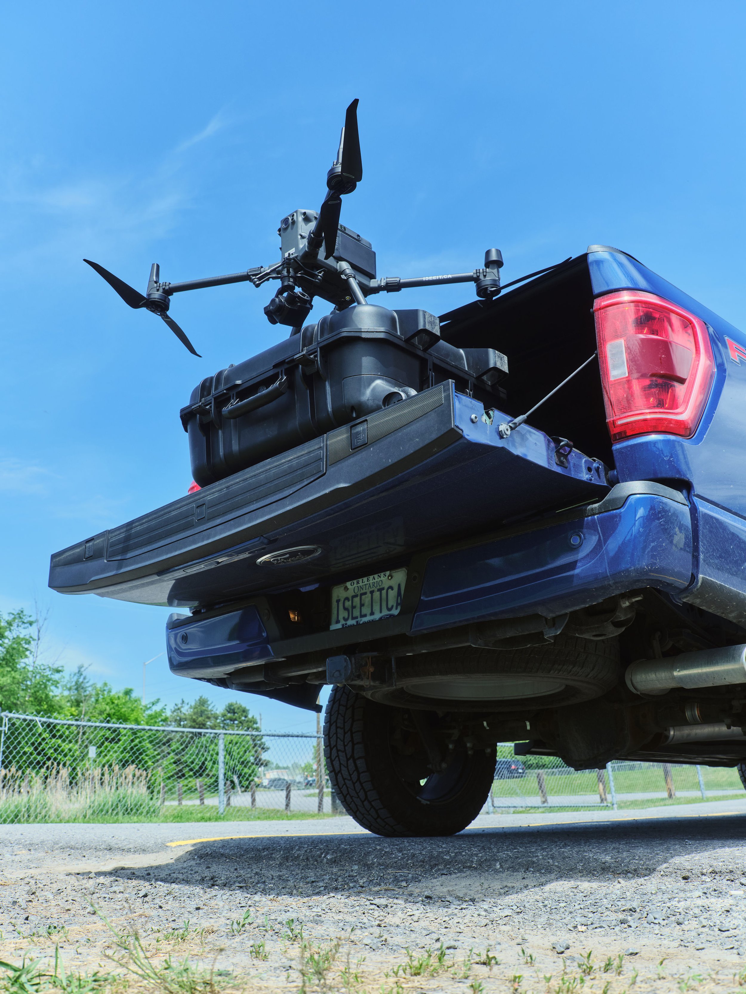 A pickup truck with a drone mounted on its cargo bed, viewed from below and slightly to the side against a clear blue sky, with a chain-link fence and trees in the background.