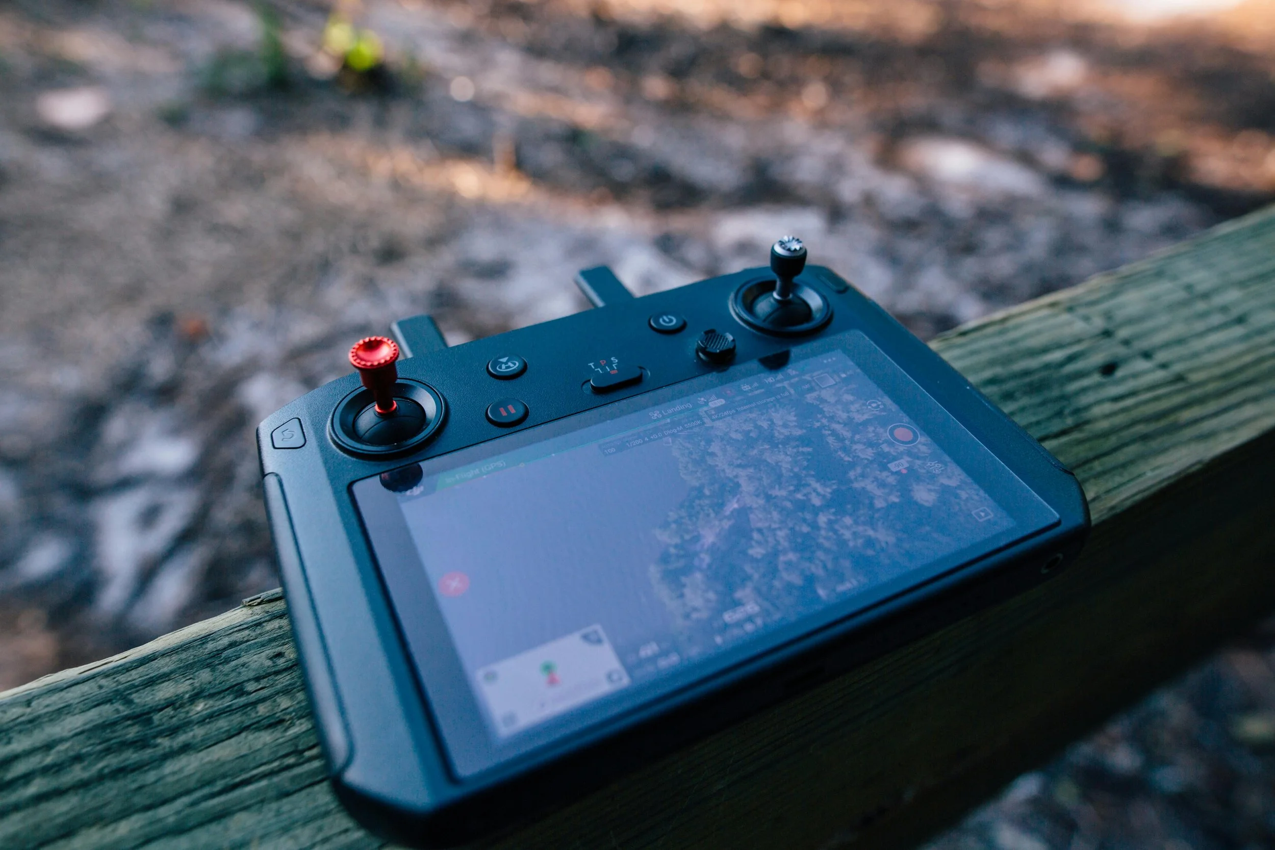 A drone remote controller with two joysticks, one with a red top and the other black, placed on a weathered wooden surface outdoors. The controller has buttons and a small touchscreen, with blurred natural background.