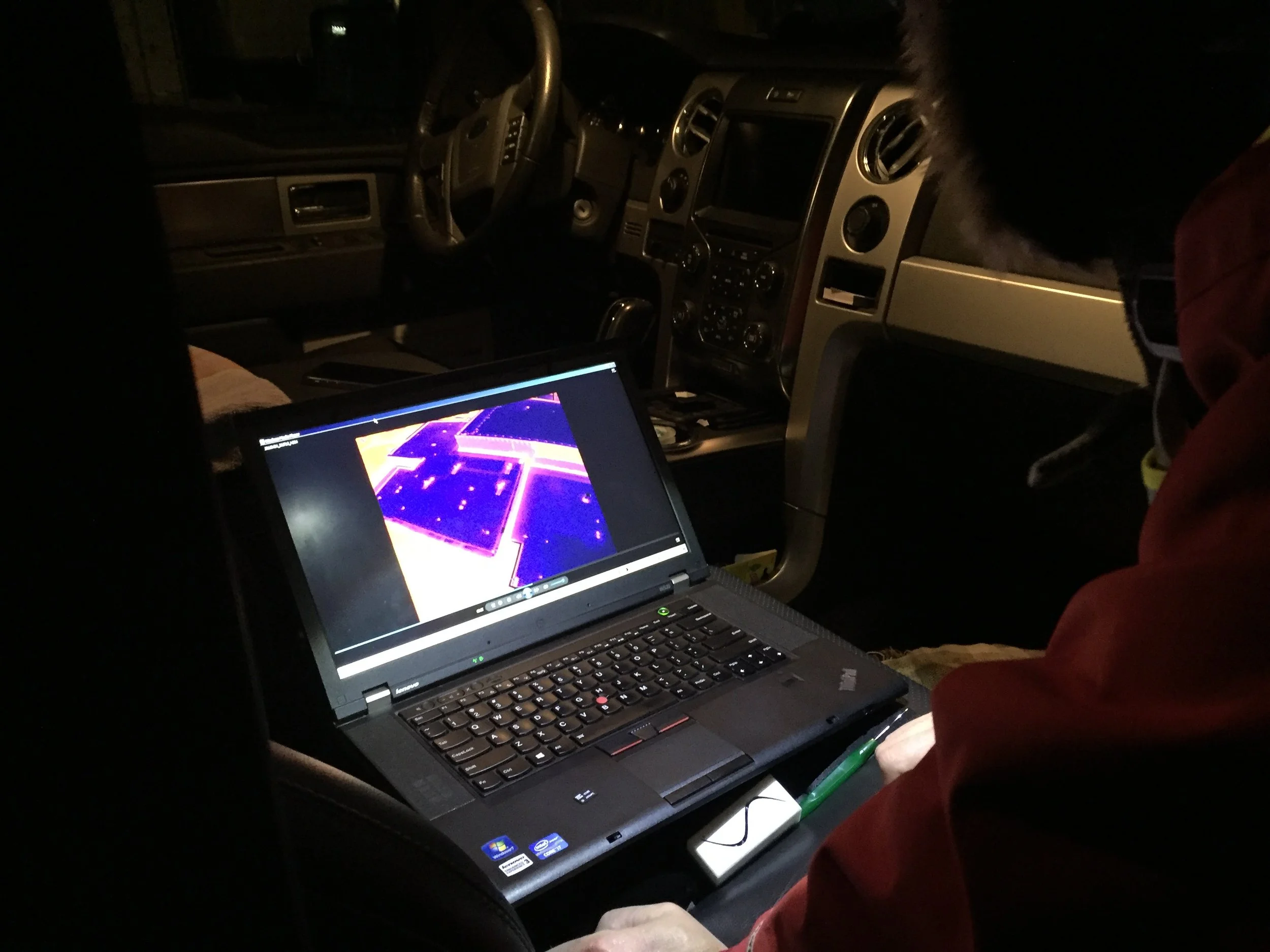 Inside a vehicle at night, a person is working on a laptop placed on their lap. The vehicle's dashboard and steering wheel are visible, and a person wearing red is seated nearby. The laptop screen displays a neon-colored design or diagram.