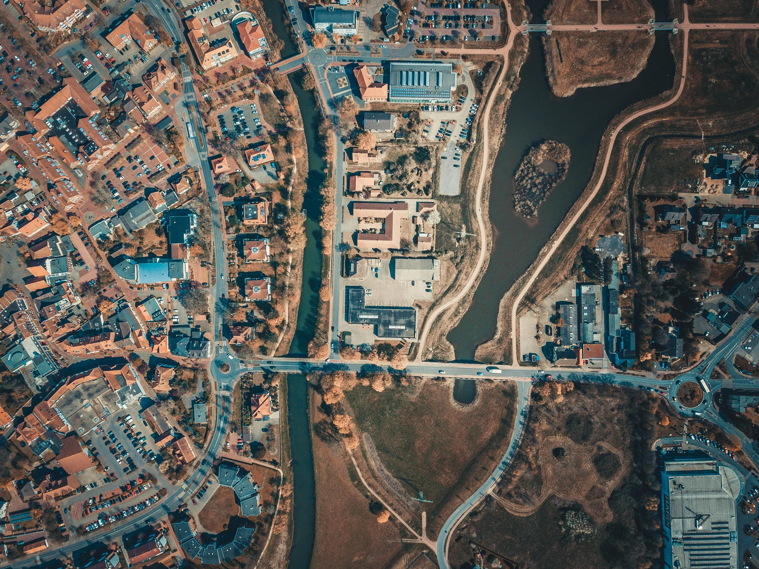 An aerial view of a suburban area with a lake, parks, roads, and residential buildings. The image shows a mix of houses, parking lots, and green spaces.