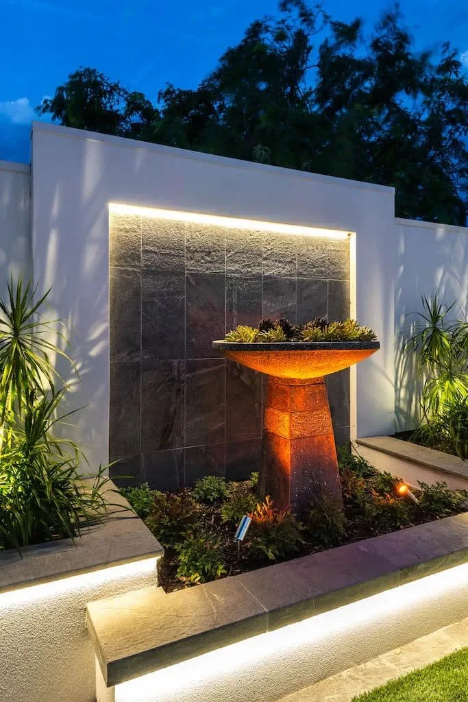 Modern outdoor wall with illuminated feature, a planter fountain with plants, surrounded by landscaping lights and greenery at dusk.