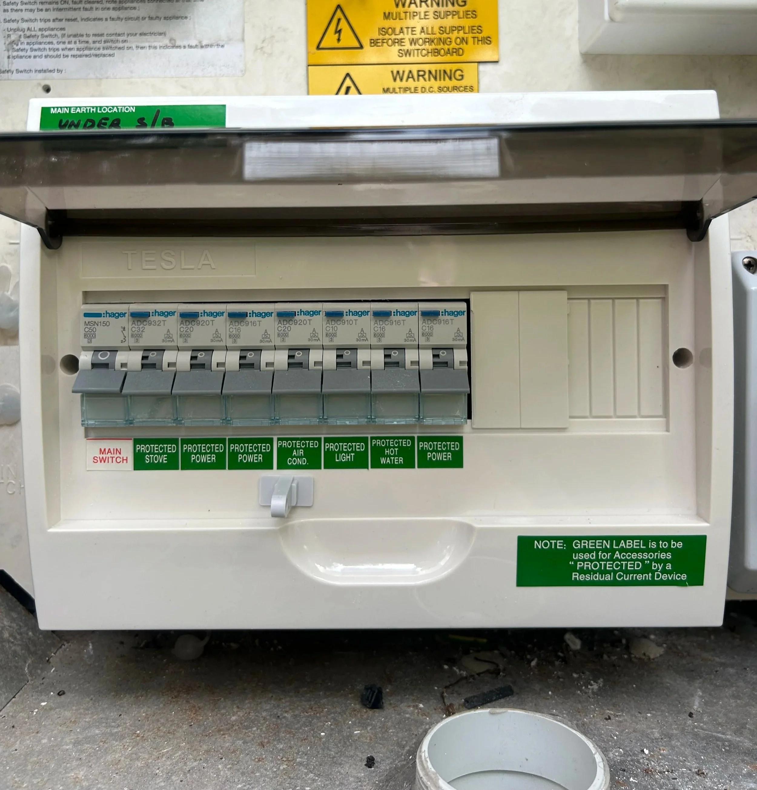 Electrical panel with circuit breakers labeled 'Tesla' and multiple protected circuits for stove, power, air conditioning, light, hot water, and power, with warning labels and instructions above.
