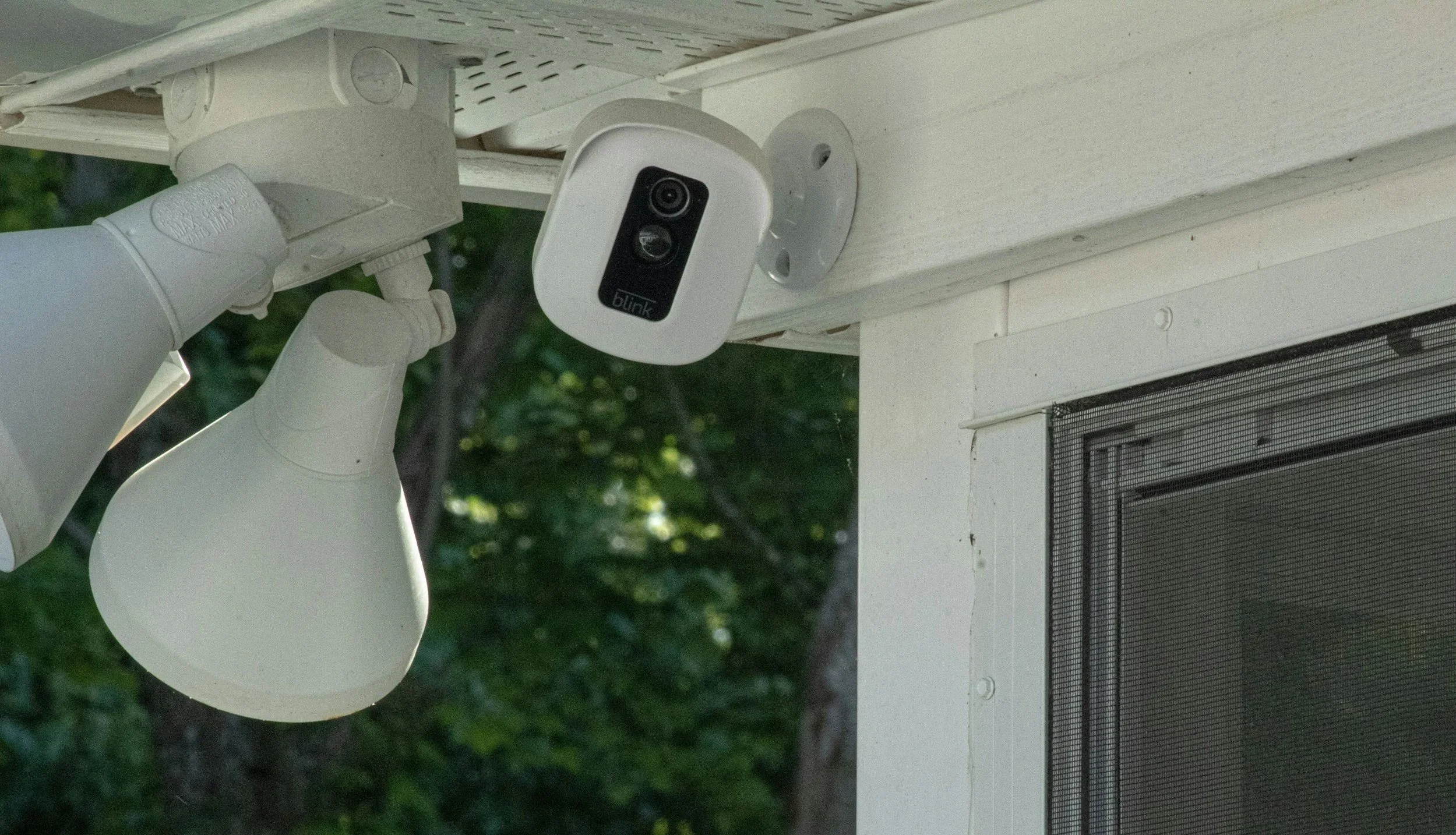Outdoor surveillance camera mounted on a white wooden porch ceiling, with two white floodlights nearby, against a background of green trees.