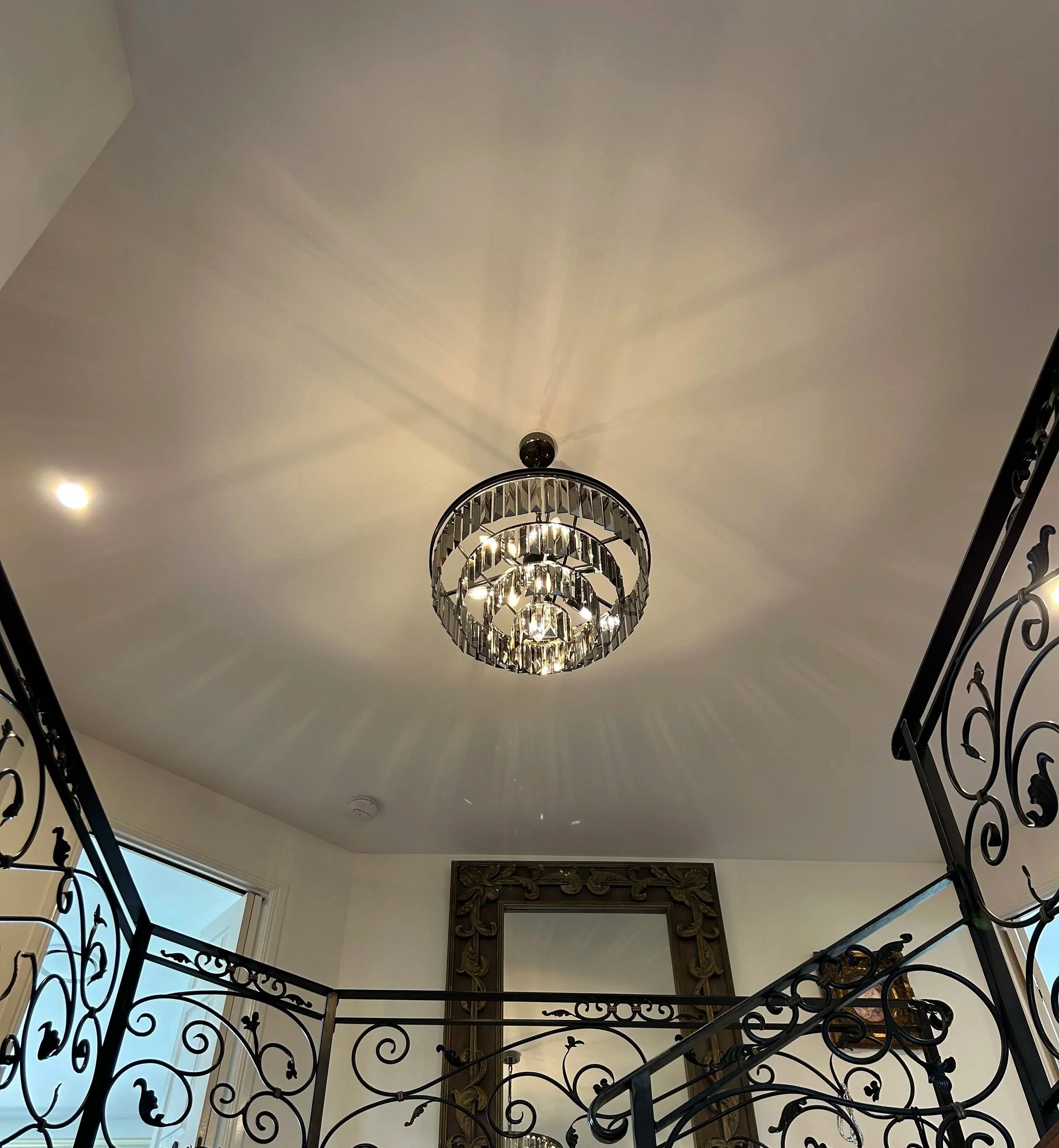 Chandelier hanging from a ceiling in a room, with decorative wrought iron railings and a mirror on the wall.