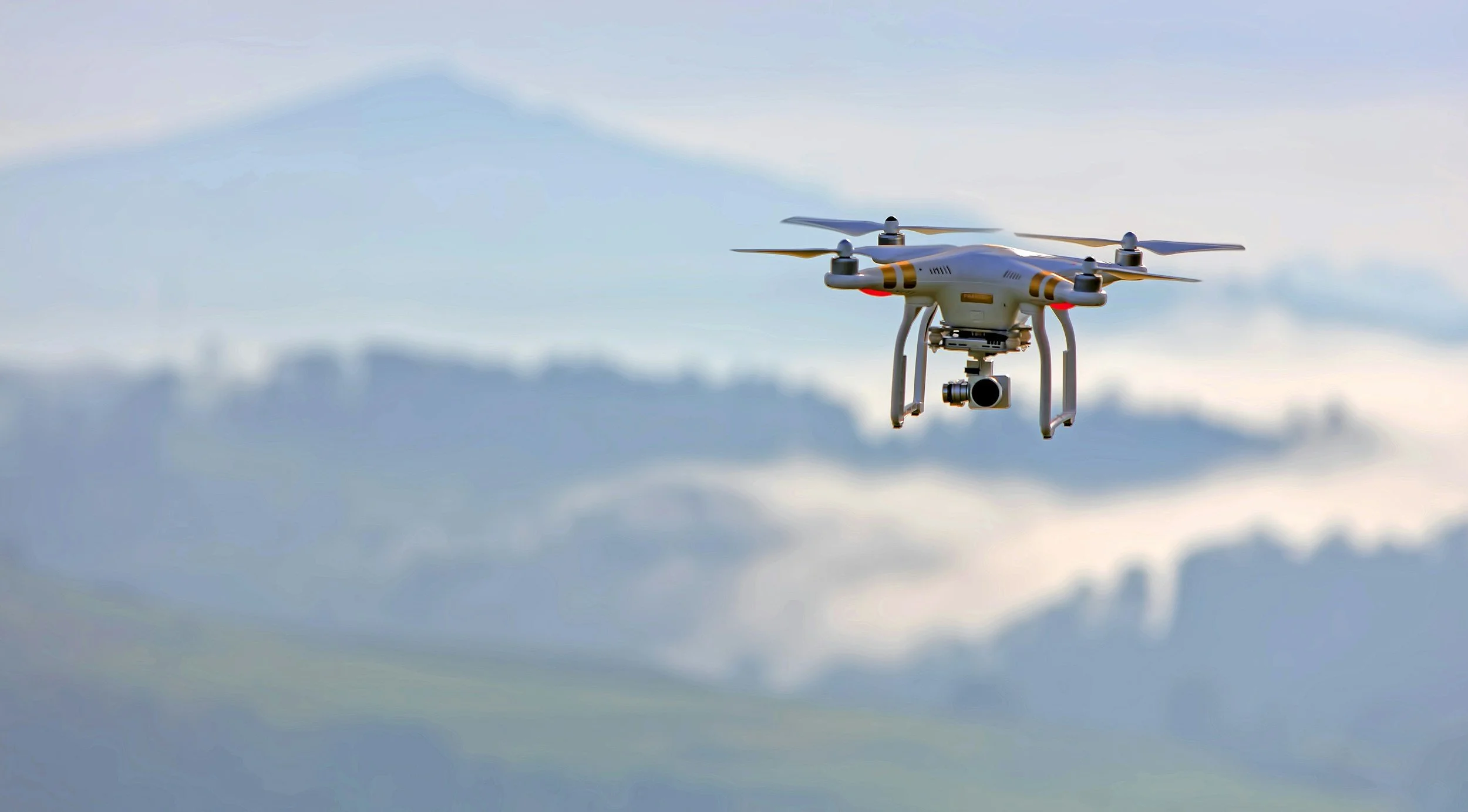 A drone with a camera attached flying in the sky with clouds and mountains in the background.