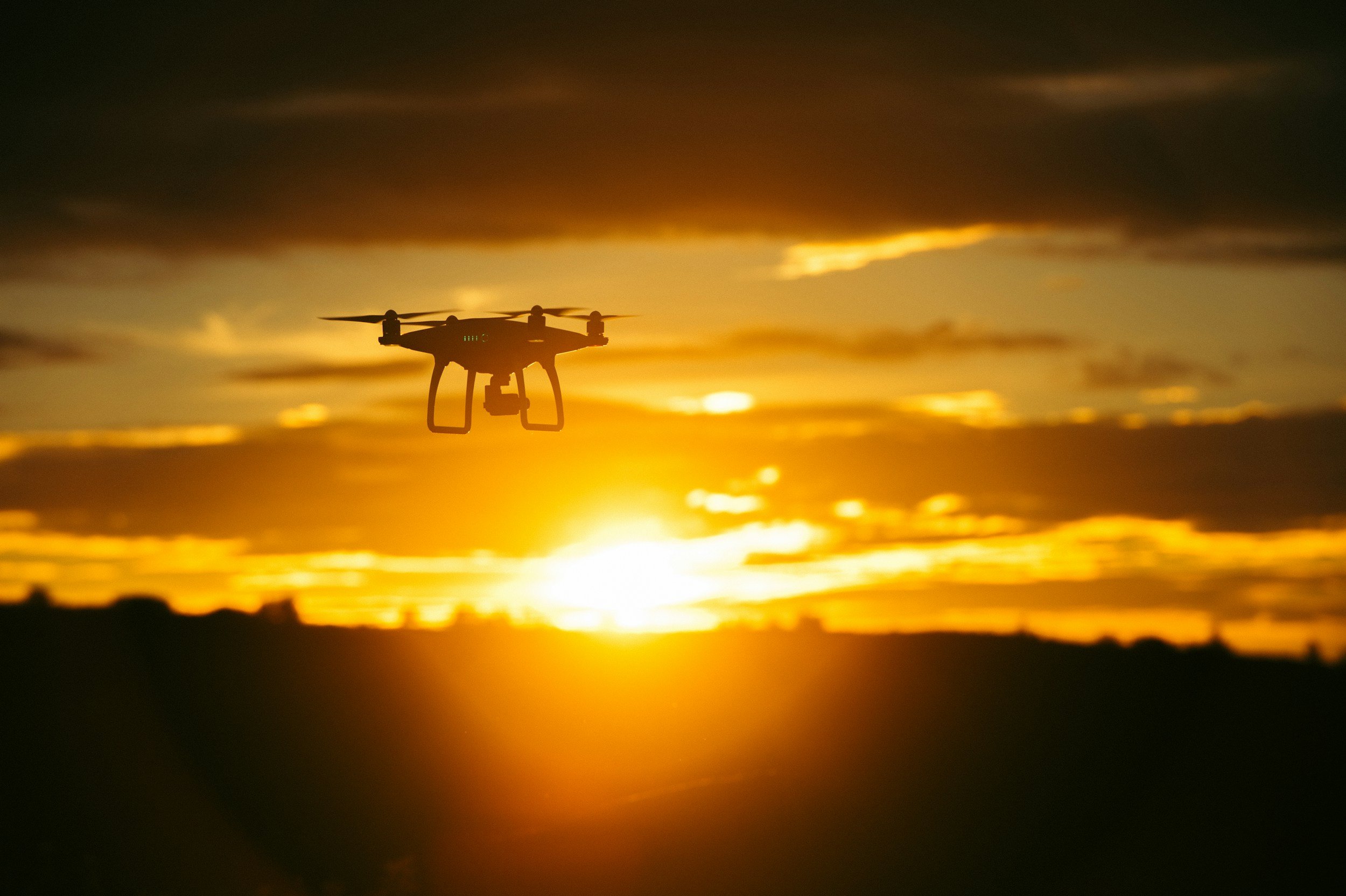 Silhouette of a drone flying against a vibrant orange sunset sky with scattered clouds.