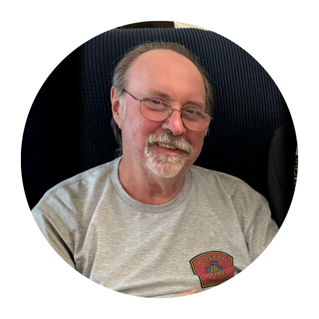 A middle-aged man with glasses and a beard, wearing a gray t-shirt with a Boston Fire Department logo, sitting against a dark background.