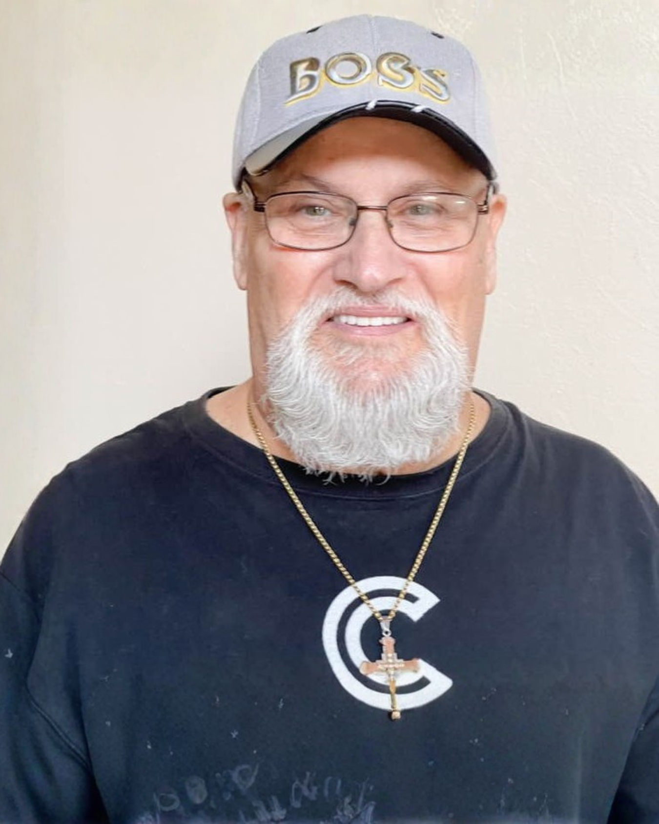 A man with glasses, a white beard, and a mustache wearing a gray cap with gold lettering that reads 'BOSS', a black t-shirt with white text, and a gold chain necklace with a cross pendant. He is standing against a plain wall, smiling at the camera.