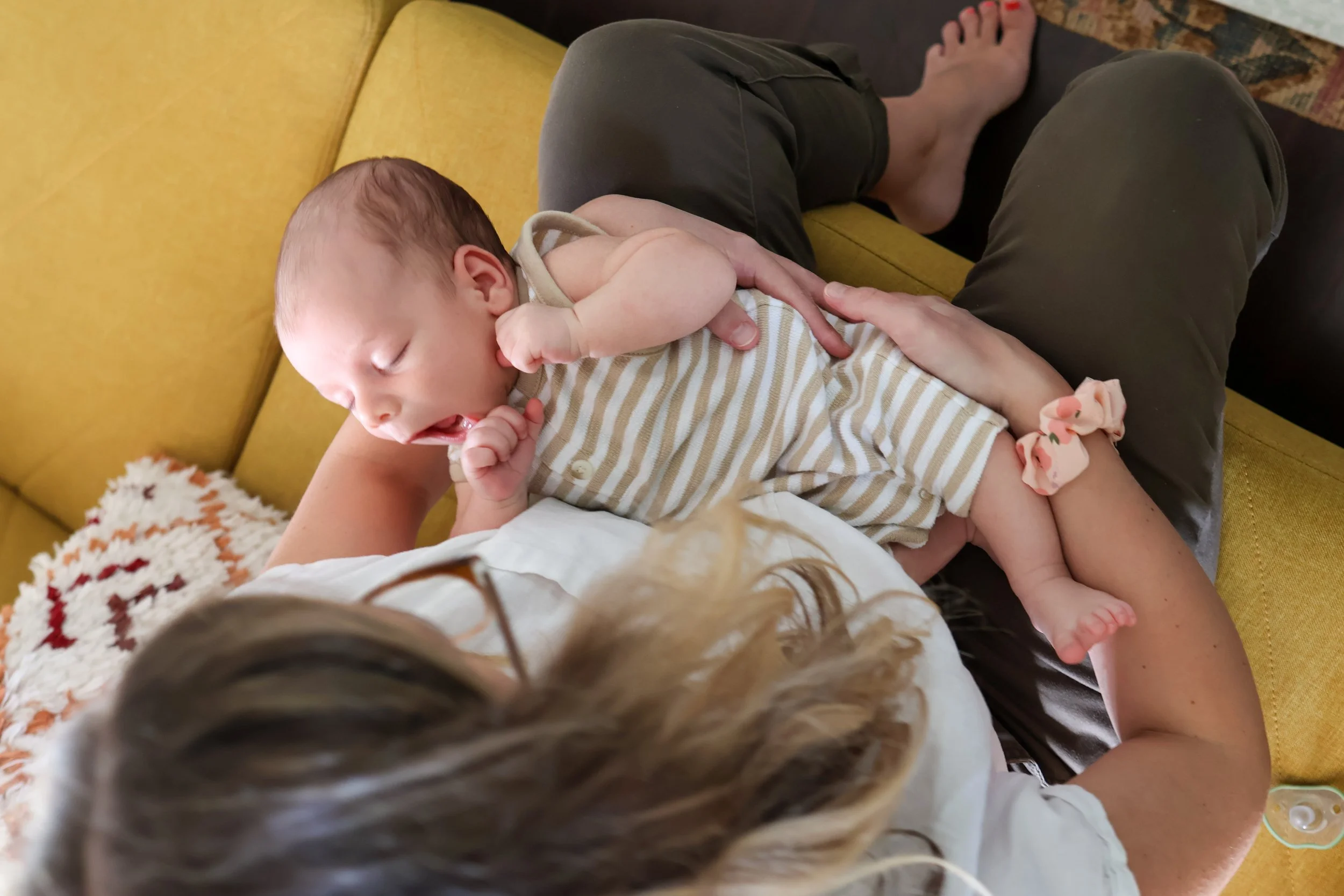 A sleeping baby laying on a woman's lap on a yellow couch, with the woman’s hand gently resting on the baby's side. The woman, with long hair and glasses, is partially visible, and a knit blanket with a red and white pattern is also seen. The scene appears to be cozy and relaxed in a living room.
